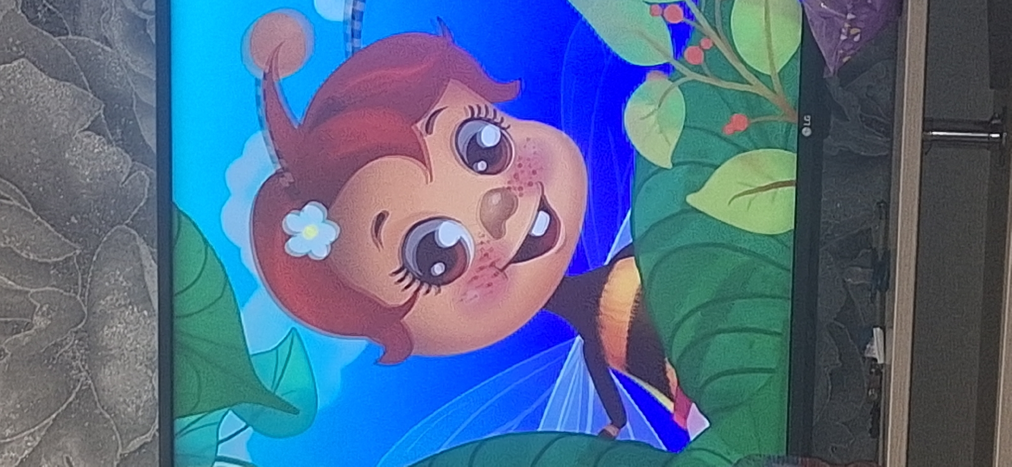 A digital illustration depicts a cartoon bee character with large eyes, rosy cheeks, and a smile. The bee has auburn hair adorned with a light blue flower and a small antennae with a pink sphere at the end. It is perched among large green leaves, against a bright blue sky with some white clouds. The lower portion of the image shows a black television screen with the LG logo visible at the bottom right. A dark shelf is below the television, with various small objects, including a white object that resembles a toy boat, and a crumpled purple plastic bag. The background above the television screen displays a textured gray pattern resembling a floral design. There is no discernible information within the image to indicate a specific location in Bila Tserkva, Ukraine.