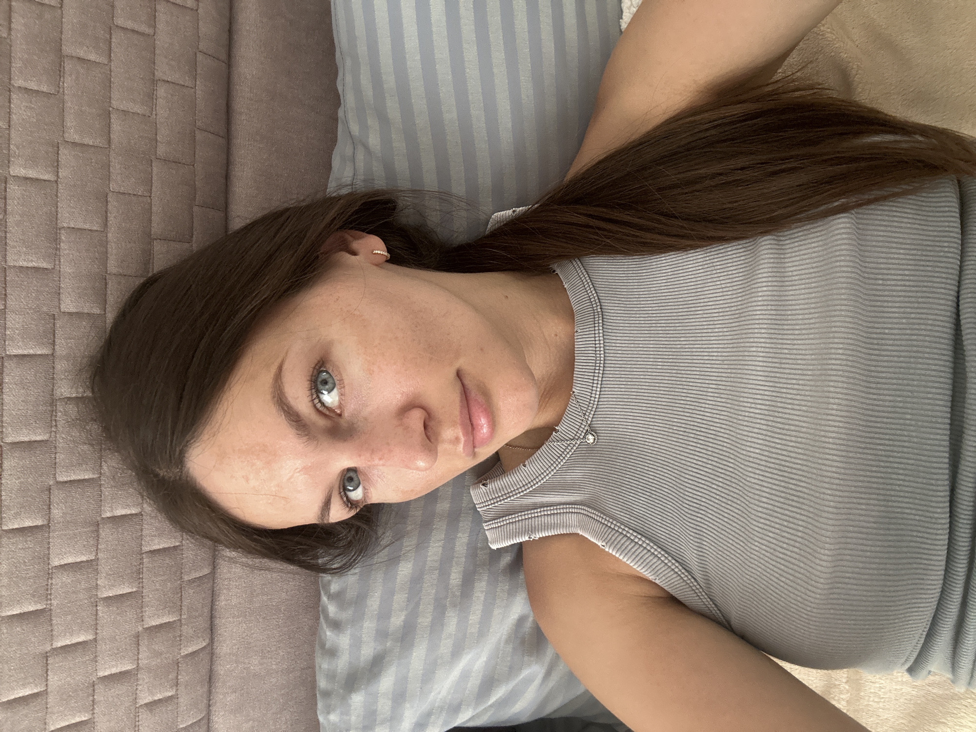 A woman is lying down on a bed or couch. Her hair is brown, and she has blue eyes. She is wearing a gray striped top. She appears to be inside a building, likely a residential space, but there is no information regarding the city of Donji Pobori, Montenegro.