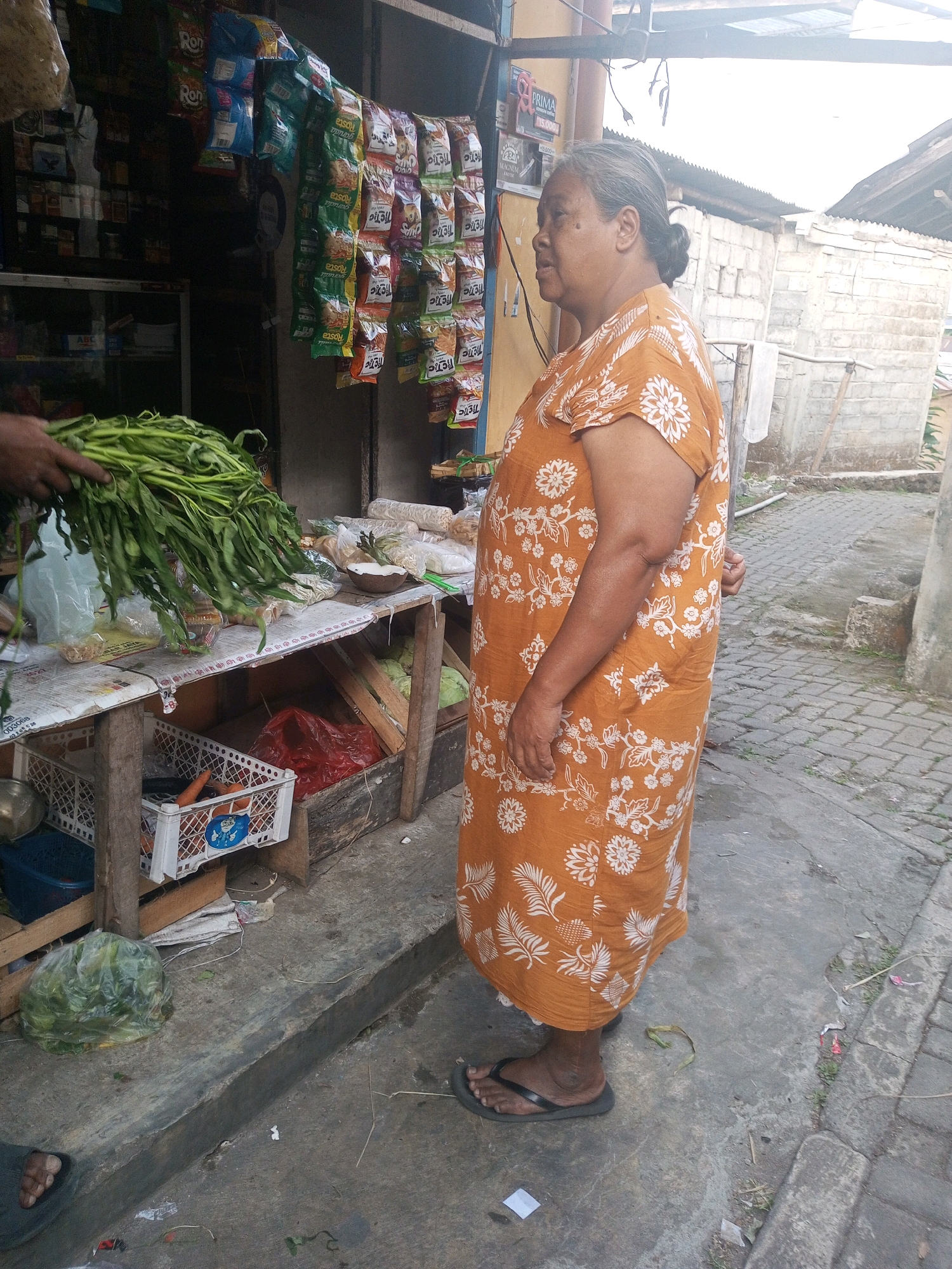An elderly woman with graying hair pulled back stands at a street-side market stall in Andongsili, Indonesia. She is wearing an orange, floral-patterned dress and dark flip-flops. She is facing left, looking towards a table laden with goods. In front of her, a bunch of green leafy vegetables, possibly water spinach, is being held by an unseen person. The stall displays various packaged snacks, some in colorful bags hanging from a rack. Below the hanging snacks, a table holds more produce and packaged items, including what appear to be dried noodles or similar goods. A white crate with carrots and other items is visible on a lower shelf. The ground is concrete, with some debris scattered around. In the background, a brick wall and a corrugated metal roof suggest a simple building structure. The scene is brightly lit by natural daylight, casting shadows on the ground.