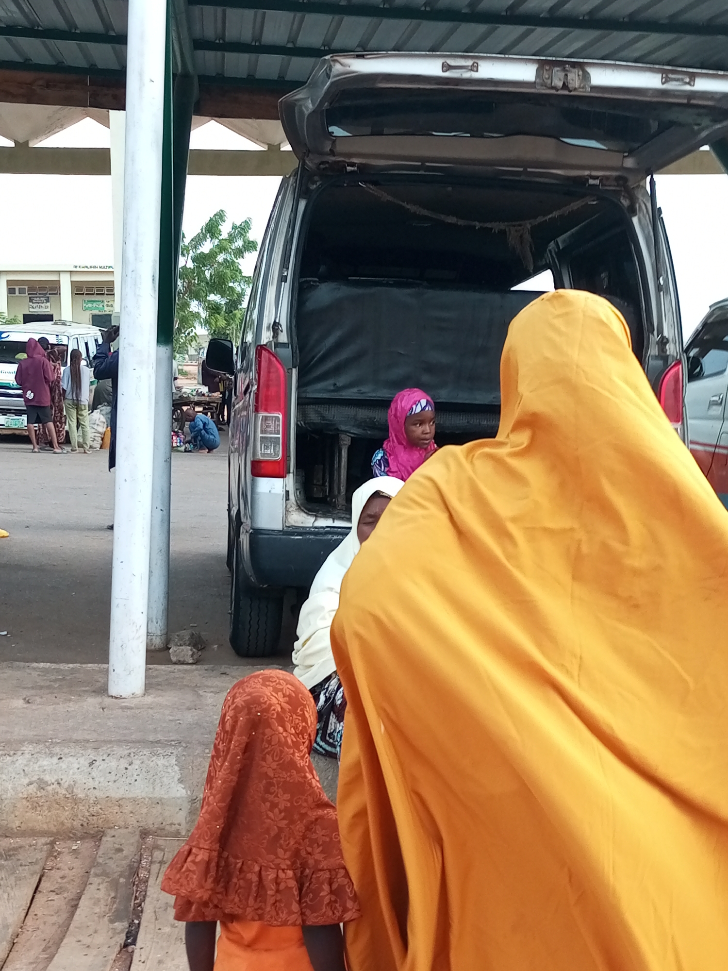 A group of people, including women and children wearing hijabs, are gathered near an open-back van in Gombe, Nigeria. The scene appears to be a marketplace or transport hub. In the foreground, a person in a bright orange hijab is facing away from the camera. To their left, a child in a red patterned hijab is also facing away. Behind them, a woman in a white hijab and a child in a pink hijab are visible inside the van. Other individuals and vehicles are present in the background, suggesting a bustling environment. The setting includes a paved ground surface, concrete pillars, and a covered structure overhead, typical of an urban setting in Nigeria.