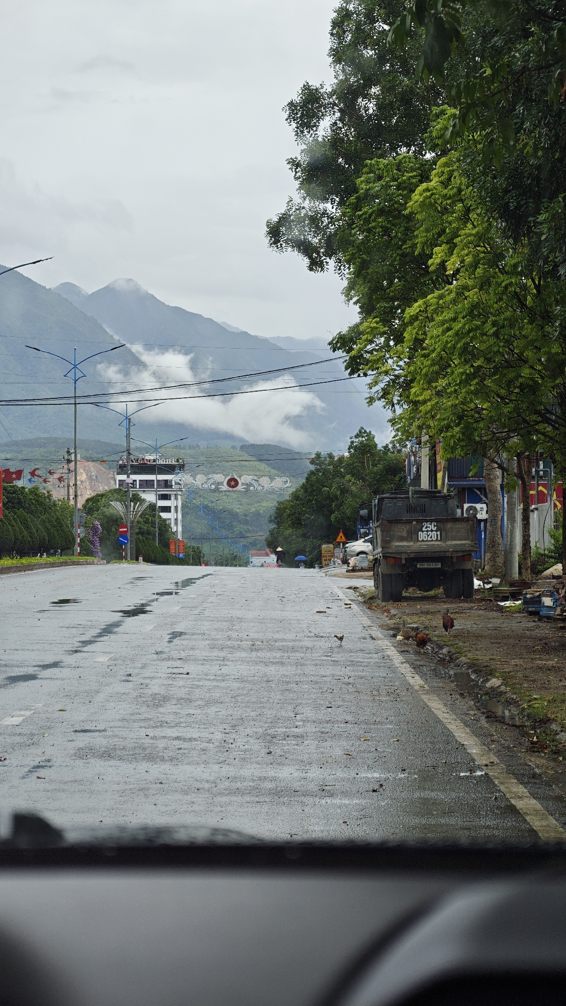 The image depicts a wet roadway in Tam Đường, Vietnam, under an overcast sky. Mountains veiled in fog are visible in the background. A building identified as "Cao Hotel" stands on the left side of the road. Power lines stretch across the scene. A truck is parked on the right shoulder of the road, next to a tree. Several chickens are near the truck. Flags are displayed on the left side of the road. The photograph appears to be taken from inside a vehicle, as part of the dashboard is visible at the bottom of the frame. A person is seen in the distance with an umbrella.