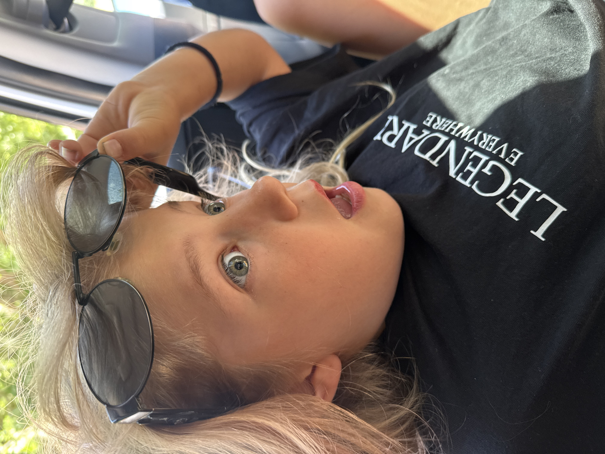 A fair-skinned child with blonde hair and green eyes is holding black sunglasses. She is lying down wearing a black t-shirt with the word "LEGENDARY" printed in white. The photograph was taken inside a vehicle, as the car doorframe is visible in the upper left corner. It is not possible to ascertain that the location is in the city of Kruče - Kërruçi, Montenegro without additional data in the image.
