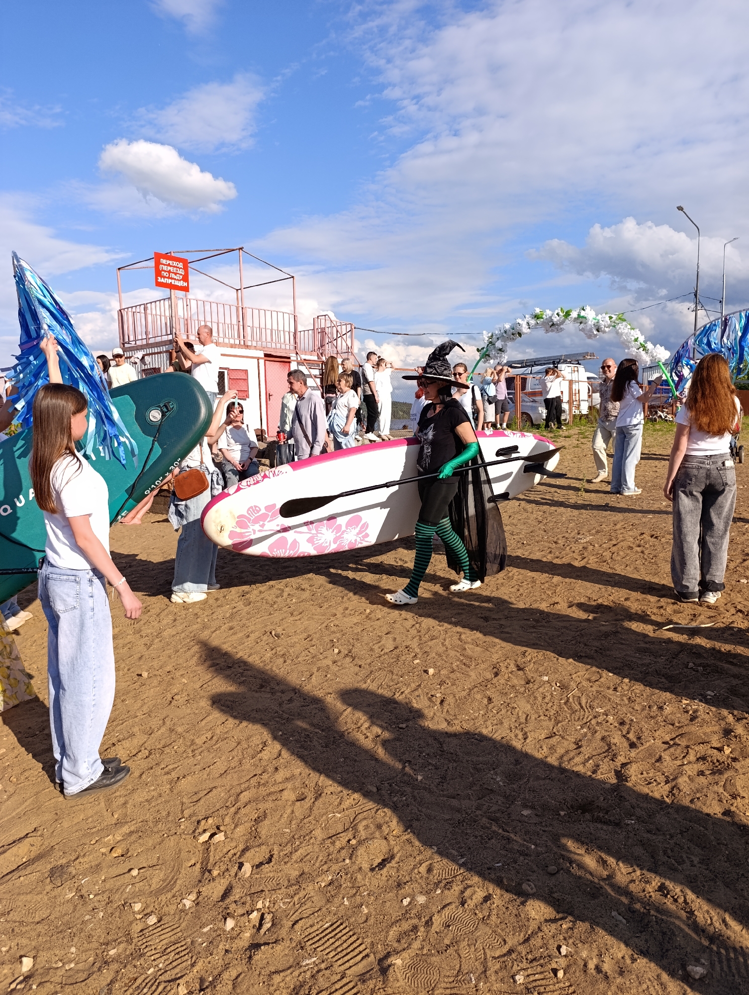 The image depicts an outdoor gathering on a sandy area, likely a beach or riverbank, on a sunny day with some clouds. A group of people are present, some holding or carrying paddleboards. In the foreground, a young woman with long brown hair, wearing a white t-shirt and light blue jeans, is looking to her left. To her right, a person in a witch's hat, black t-shirt, green striped tights, and black cape is carrying a pink and white paddleboard. Other individuals are visible in the background, some taking pictures with their phones.
Behind the people, there is a temporary structure with a red roof and railings, and a sign with Russian text that appears to be a warning. Further in the background, a floral archway is set up, suggesting a celebration or event. The overall atmosphere appears to be festive and casual. The lighting indicates it is daytime, likely late afternoon given the long shadows cast on the sand. The presence of paddleboards and the beach-like setting suggest a recreational activity. The location context provided is Chaikovsky, Russia.