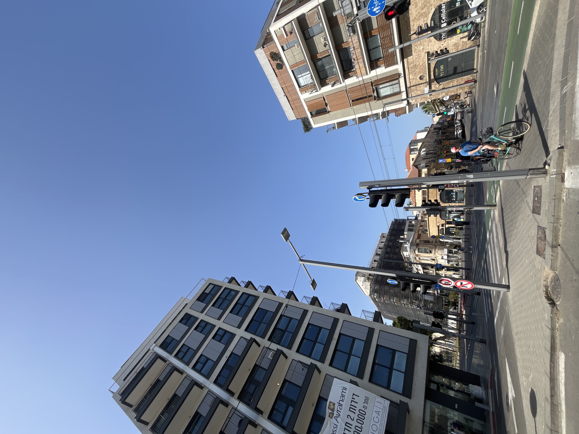 The image captures a street view in Tel Aviv, Israel. In the foreground, a person rides a bicycle on a green bike lane along a crosswalk, as the traffic light facing them appears red. In the backdrop, modern buildings rise against a clear blue sky. One building has an advertisement promoting the sales of 2-room apartments. A traffic light pole and signs are also present, featuring directions and restrictions for vehicular traffic.