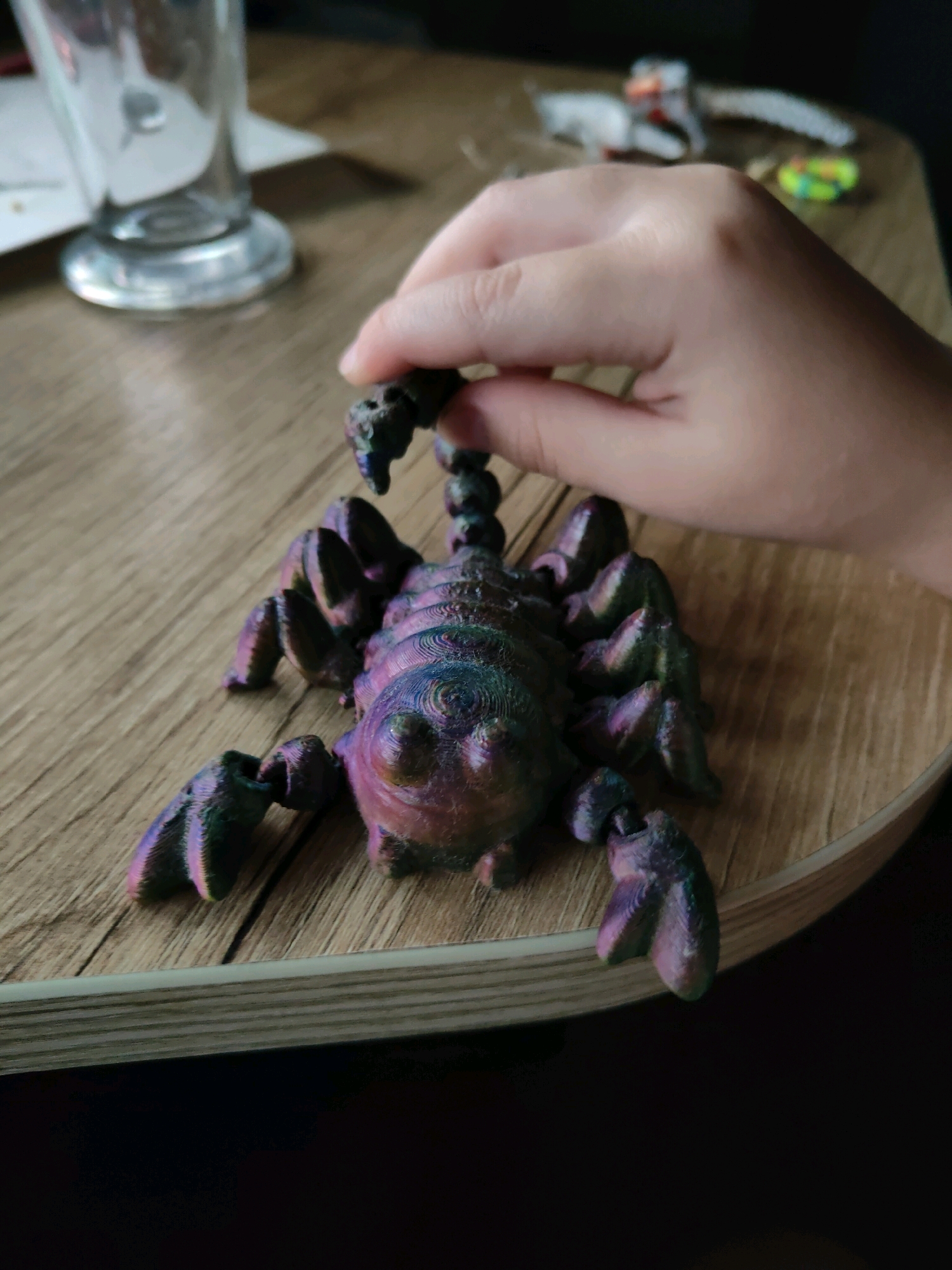 In Homyel, Belarus, a close-up shot focuses on a person's hand holding a 3D-printed scorpion toy with a metallic, iridescent finish, displaying hues of purple, green, and blue. The scorpion is positioned on a wooden surface, its articulated legs and segmented tail clearly visible. To the left, the base of a glass is partially in view, and in the background, out-of-focus objects suggest a table setting. The lighting creates a warm ambiance, highlighting the texture and sheen of the toy.