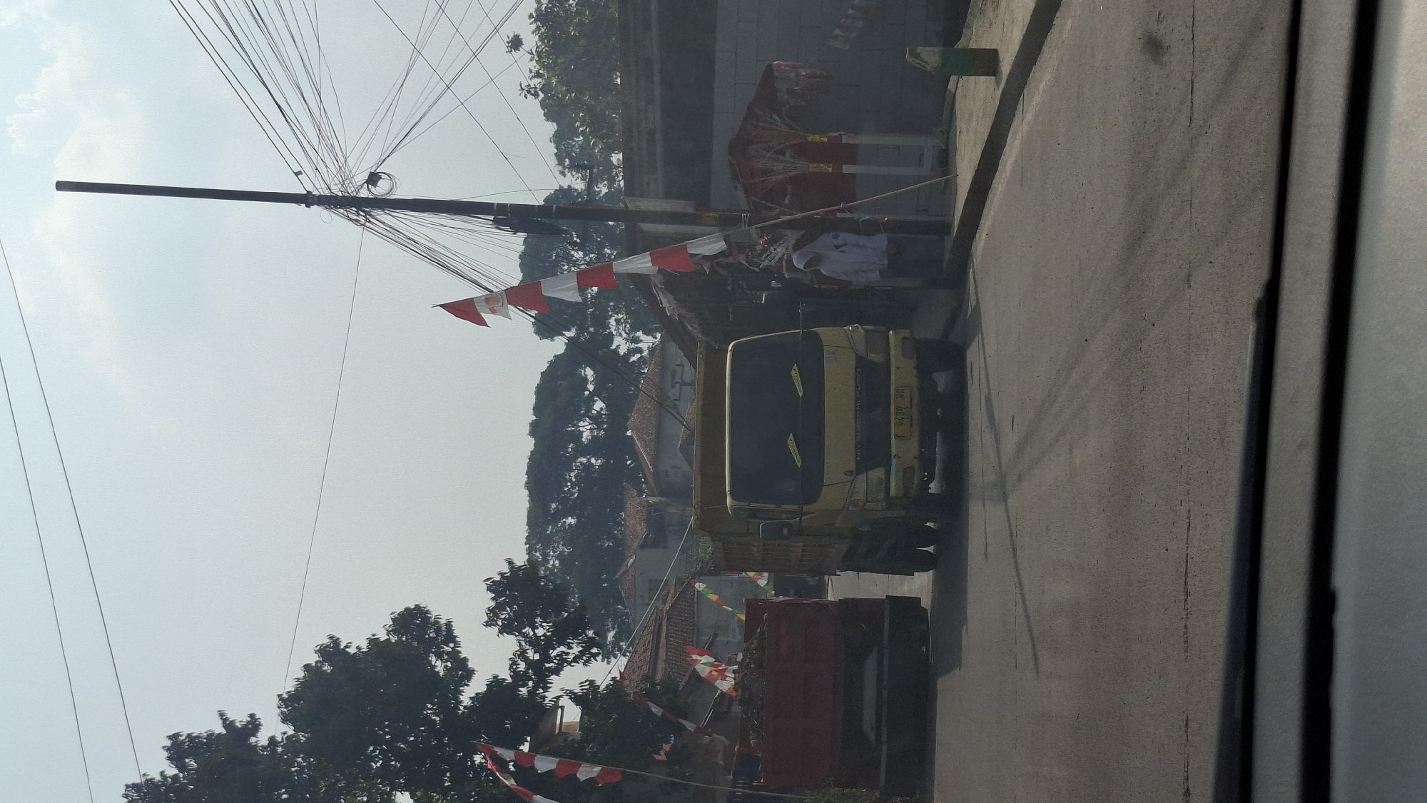 An outdoor, daytime, eye-level medium shot captures a street scene in Cilaku, Indonesia, with Indonesian flags decorating the area. Two dump trucks are visible on the asphalt road. The truck on the left is red and filled with dark debris. The truck on the right is yellow and appears to be a Mitsubishi Fuso, with license plate number F 9439 WA. A person wearing a white hijab and white jacket stands on the right side of the road next to a utility pole adorned with a string of Indonesian flags. The background features trees, houses with red tile roofs, and a grey wall. Overhead, a network of electrical wires and a utility pole dominate the sky. The road shows tire tracks and a few scattered pieces of debris. The image appears to be taken from inside a vehicle, indicated by the dark border at the bottom of the frame.