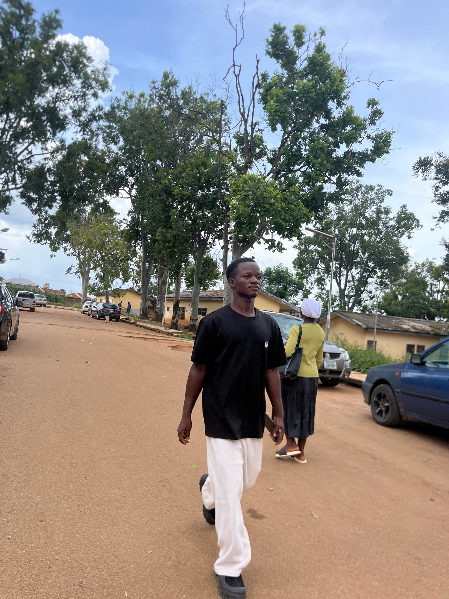 The image shows a street scene in the city of Rafin Par, Nigeria. A young man in a black t-shirt and white pants is walking towards the camera. Behind him, a woman in a yellow top and black skirt, with a white headscarf, is walking away. Several parked cars are visible on the side of the road, and some low-rise buildings with yellow walls line the street in the background. Tall trees with green foliage are scattered throughout the scene, and the sky is partly cloudy. The road appears to be paved with reddish-brown earth.