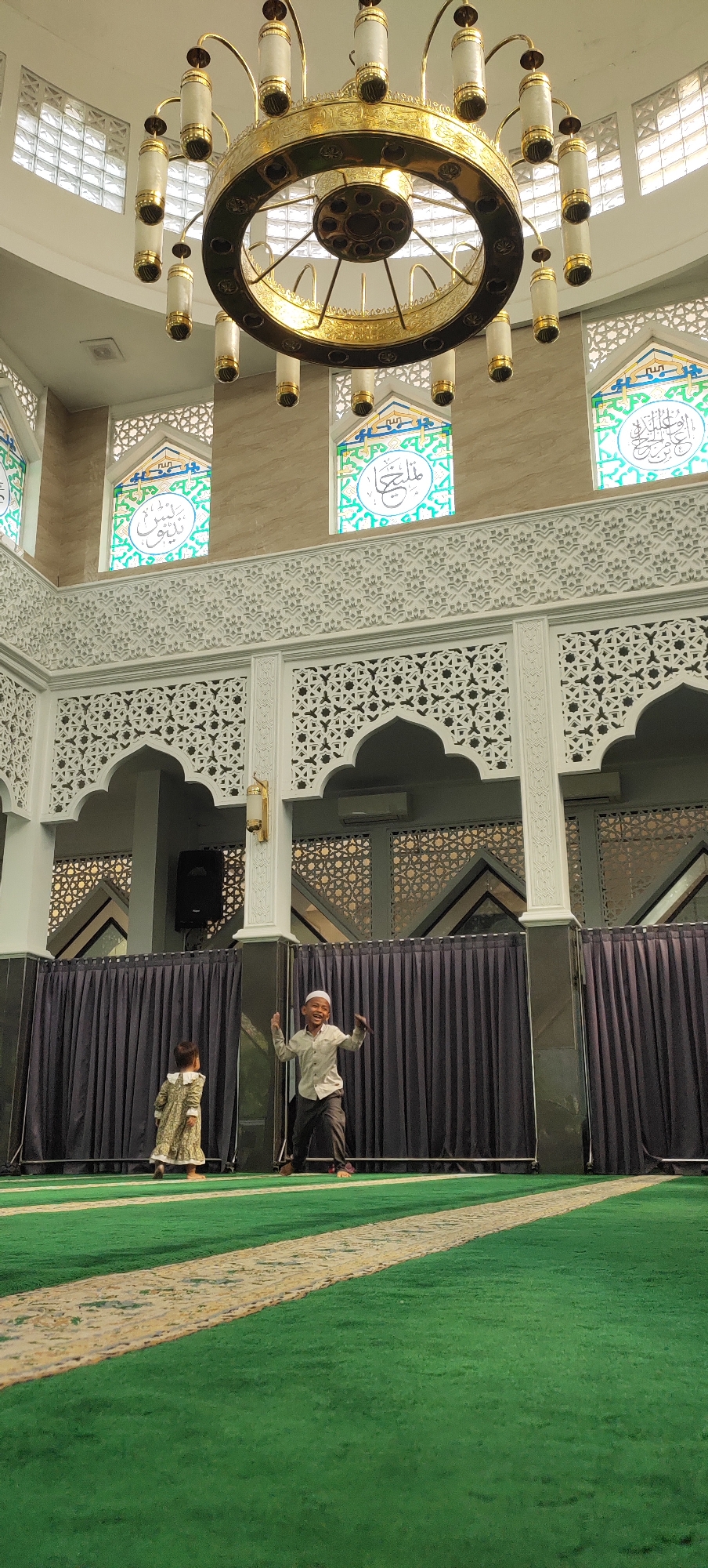 A wide-angle, eye-level, vertical shot captures the interior of a mosque in Jatirejo, Indonesia. The scene is dominated by a large, ornate golden chandelier hanging from the ceiling, with multiple cylindrical lights. Below, two children are visible on a green carpeted floor. To the right, a young boy wearing a white cap and a light-colored shirt and dark pants is jumping with joy, arms outstretched. To the left, a young girl in a patterned dress faces away from the camera, walking towards the boy. The background features dark curtains, ornate white architectural details with geometric patterns, and stained-glass windows with Arabic calligraphy. The lighting is bright and diffused, creating a well-lit environment.