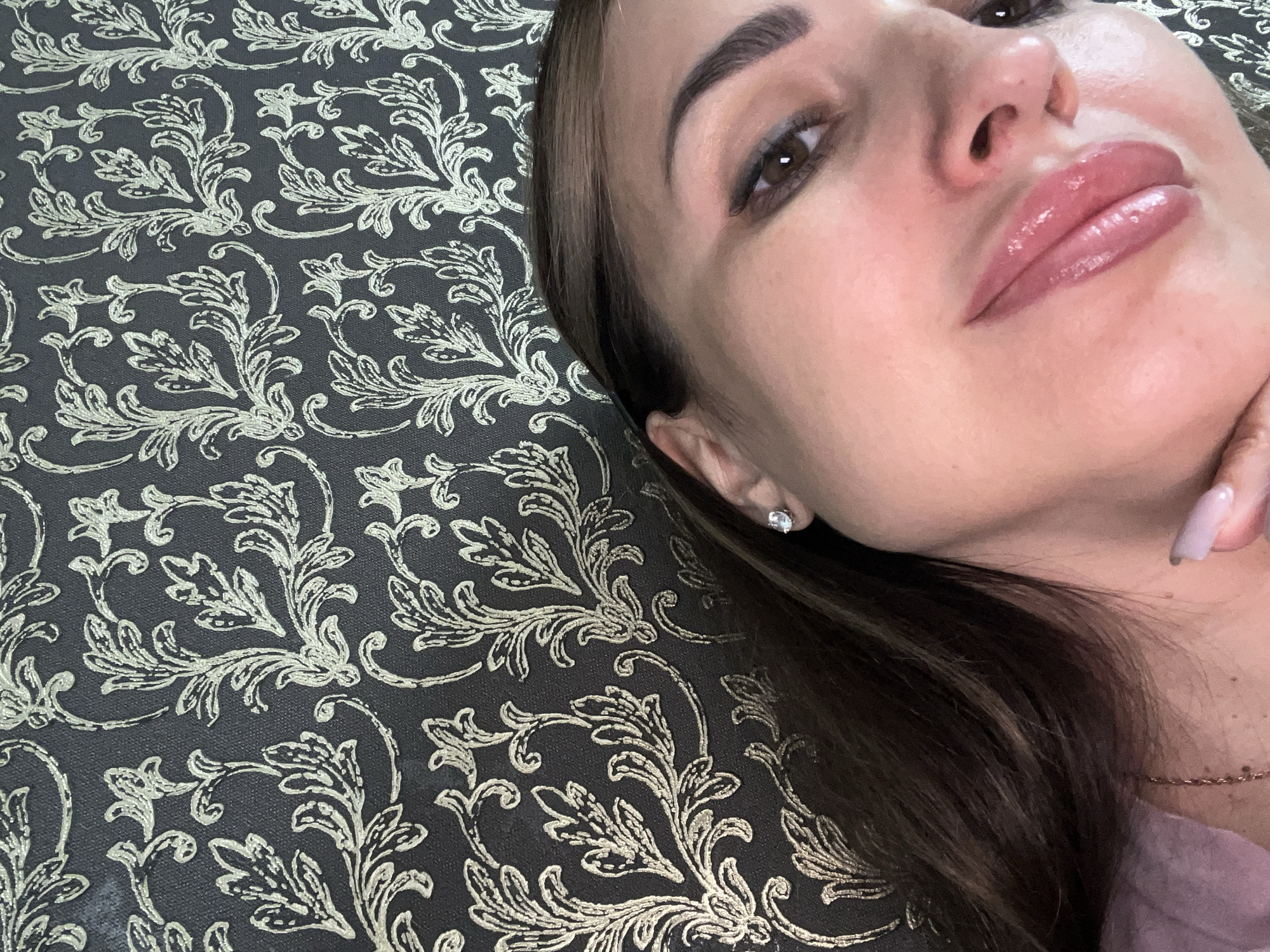 A close-up, high-angle shot shows a woman's face from the nose up against a dark patterned wallpaper. The woman has brown hair, a small diamond stud earring in her left ear, and is wearing light pink lipstick. Her lips are slightly parted and glossy. Her right eye is visible, with dark eyeliner and mascara. The wallpaper features a repeating gold floral pattern on a dark background. The scene appears to be indoors, likely in a home or studio.