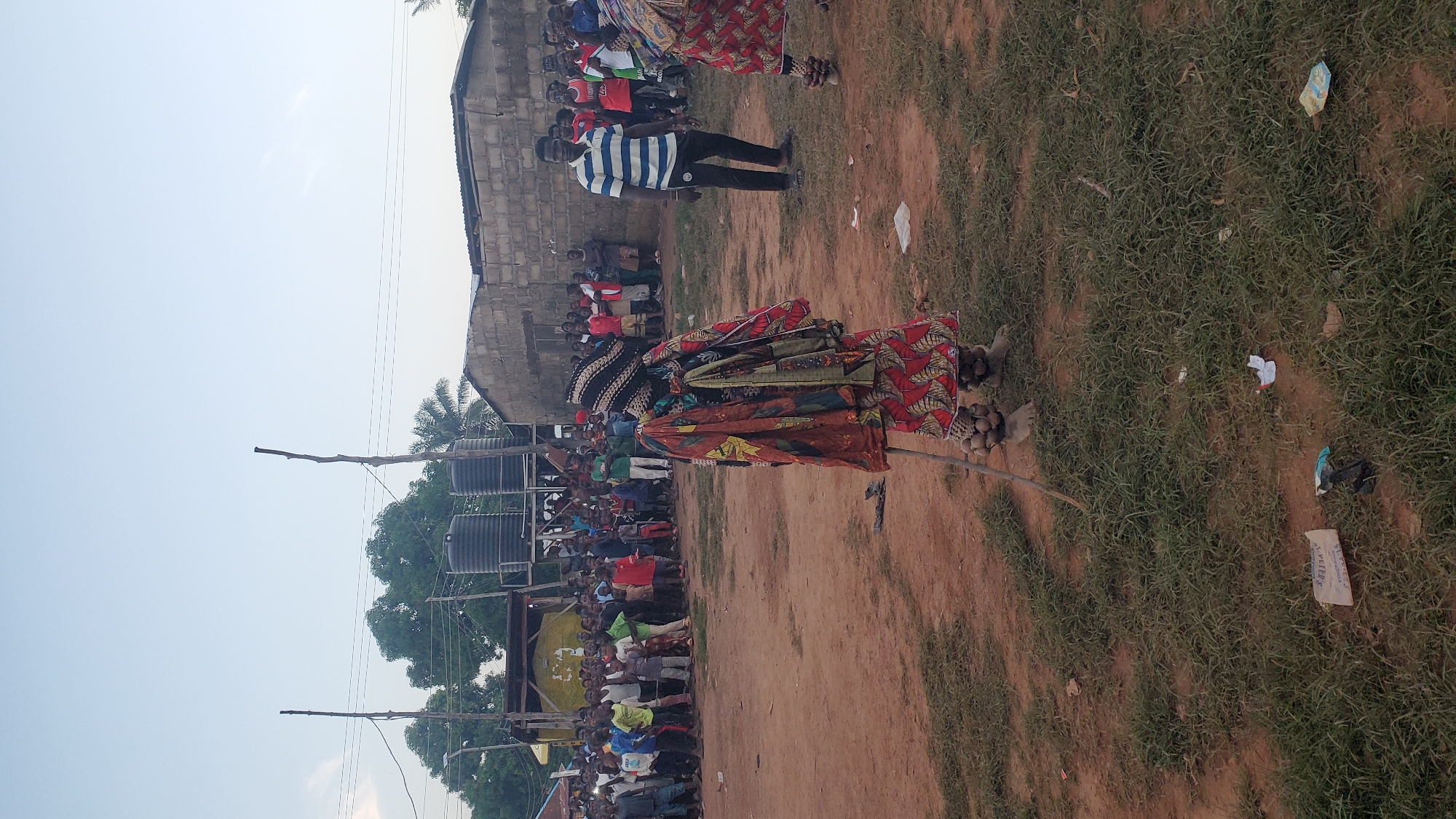 An outdoor daytime scene in Akwanga, Nigeria, depicts a festive gathering with a person in traditional costume as the central focus. This individual is adorned in a vibrant, patterned cloth ensemble, including a headdress and ankle adornments. The person holds a wooden staff and appears to be either dancing or posing for a photograph.
Surrounding the costumed individual is a crowd of onlookers, predominantly men and boys, with a smaller number of women. They are dressed in casual attire, with some wearing t-shirts and shorts, others in more traditional garments. The background includes a partially constructed brick building and what appear to be water tanks on a raised platform, suggesting a semi-urban or rural setting. The ground is a mix of dry, reddish earth and sparse green grass, with some scattered debris. The sky is clear and bright, indicating fair weather. The overall atmosphere appears to be one of community celebration or cultural event.