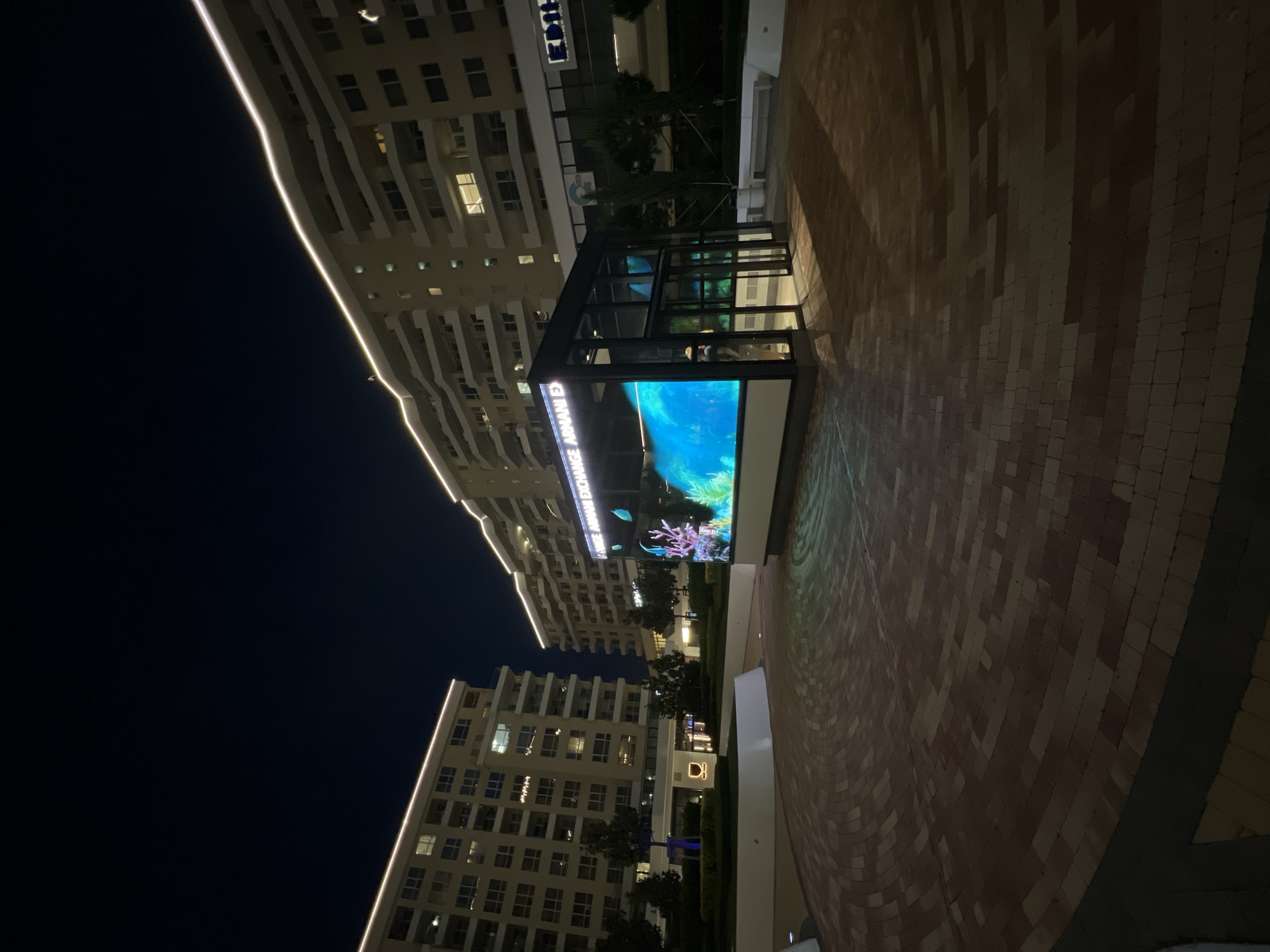 The image depicts an urban outdoor area at night in Burgas, Bulgaria, characterized by modern architecture and digital displays. Two multi-story buildings, likely residential or mixed-use, frame the scene, featuring light-colored facades and numerous balconies. Both buildings are illuminated along their rooflines with continuous white strip lighting, creating a distinct glowing perimeter against the dark night sky. Some windows in the buildings display interior lighting.
In the foreground, a large circular plaza paved with reddish-brown and tan bricks in concentric patterns occupies the majority of the lower frame. Prominently positioned in the center-right of this plaza is a modern, glass-and-metal structure. One side of this structure features a large, vivid digital display exhibiting an aquatic scene with brightly colored corals and blue water, suggesting an interactive or decorative element. Text visible on the top of this display reads "CHANGE ARMANI EXCH", indicating "Armani Exchange". The blue and green light from this digital screen casts reflections onto the paved ground directly in front of it.
Inside the glass structure, to the right of the main digital screen, an individual is partially visible, facing away from the camera, their features indistinct due to distance and lighting. The surrounding area includes landscaped sections with small trees and shrubs, some within raised white planters. One tree on the left side of the frame is illuminated at its base by a blue light. The overall environment suggests a contemporary commercial or leisure complex under a clear, dark night sky.