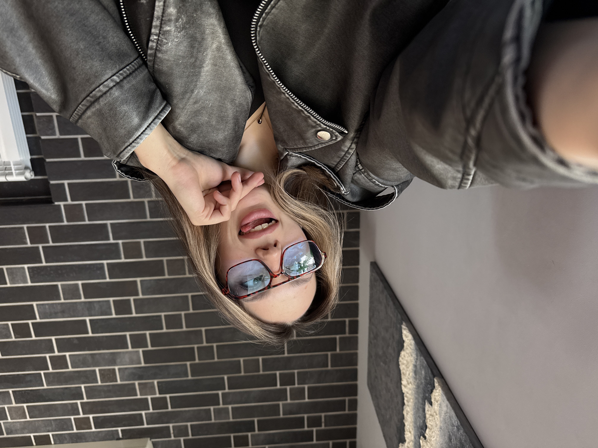 A person is holding a camera to take a selfie in a room. The person is wearing red eyeglasses, a black shirt, and a gray leather jacket. They have their hand near their face and are sticking their tongue out. The room has a dark gray brick wall, a white radiator, and a gray wall with a framed artwork. The location is Unknown, Unknown.