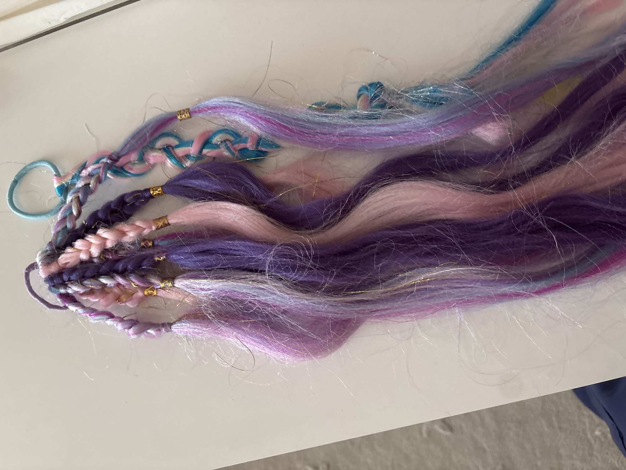The image displays a close-up, top-down view of several braided hair extensions, arranged on a white surface. The extensions are multi-colored, featuring shades of pink, purple, light blue, and white, with some strands of gold tinsel woven throughout. Each braid is secured at the top with a small, gold bead or crimp. One of the braids is attached to a light blue elastic hair tie. The surface appears to be a table or countertop. The background shows a textured floor and a portion of what might be a dark blue garment. There are no discernible landmarks or clues indicating a specific location in Dubai, United Arab Emirates.