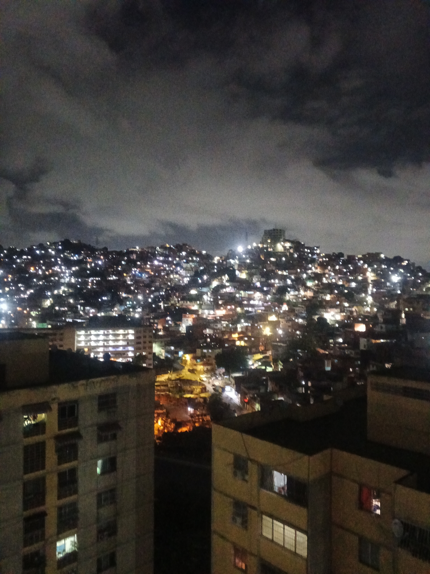 A night-time, low-angle long shot depicts a section of Caracas, Venezuela. The foreground shows the flat roofs and windows of residential high-rise buildings, with some windows lit. Behind these buildings, a hill is densely packed with houses, their lights creating a constellation-like effect. At the crest of this hill, a larger, more prominent building or structure is visible. The sky is dark and covered with clouds.