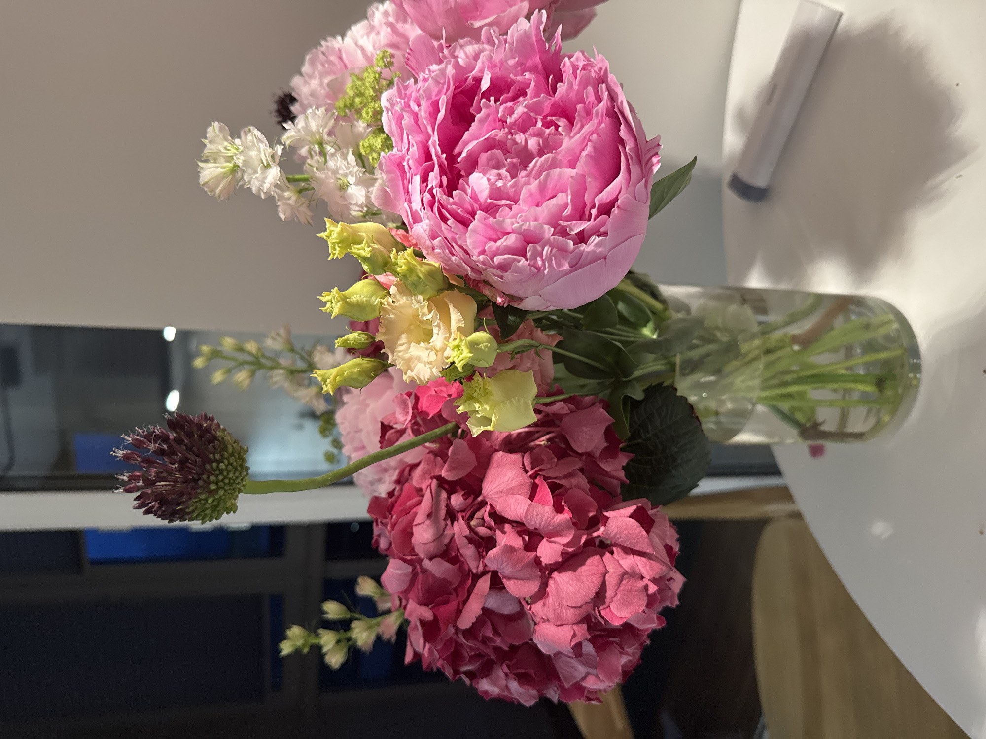 A vase of pink and white flowers sits on a white table in front of a window. The flowers include hydrangeas, peonies, lisianthus, and an ornamental onion. The window is dark, with some blue light visible, suggesting it is nighttime or dusk. The table is round and white. To the right of the vase, a cylindrical object with a grey tip lies on the table. A blurred background shows a dimly lit interior and a glimpse of a wooden chair. The overall lighting is soft, with some shadows cast on the table by the vase and flowers. The scene appears to be indoors, possibly a home or a commercial establishment.