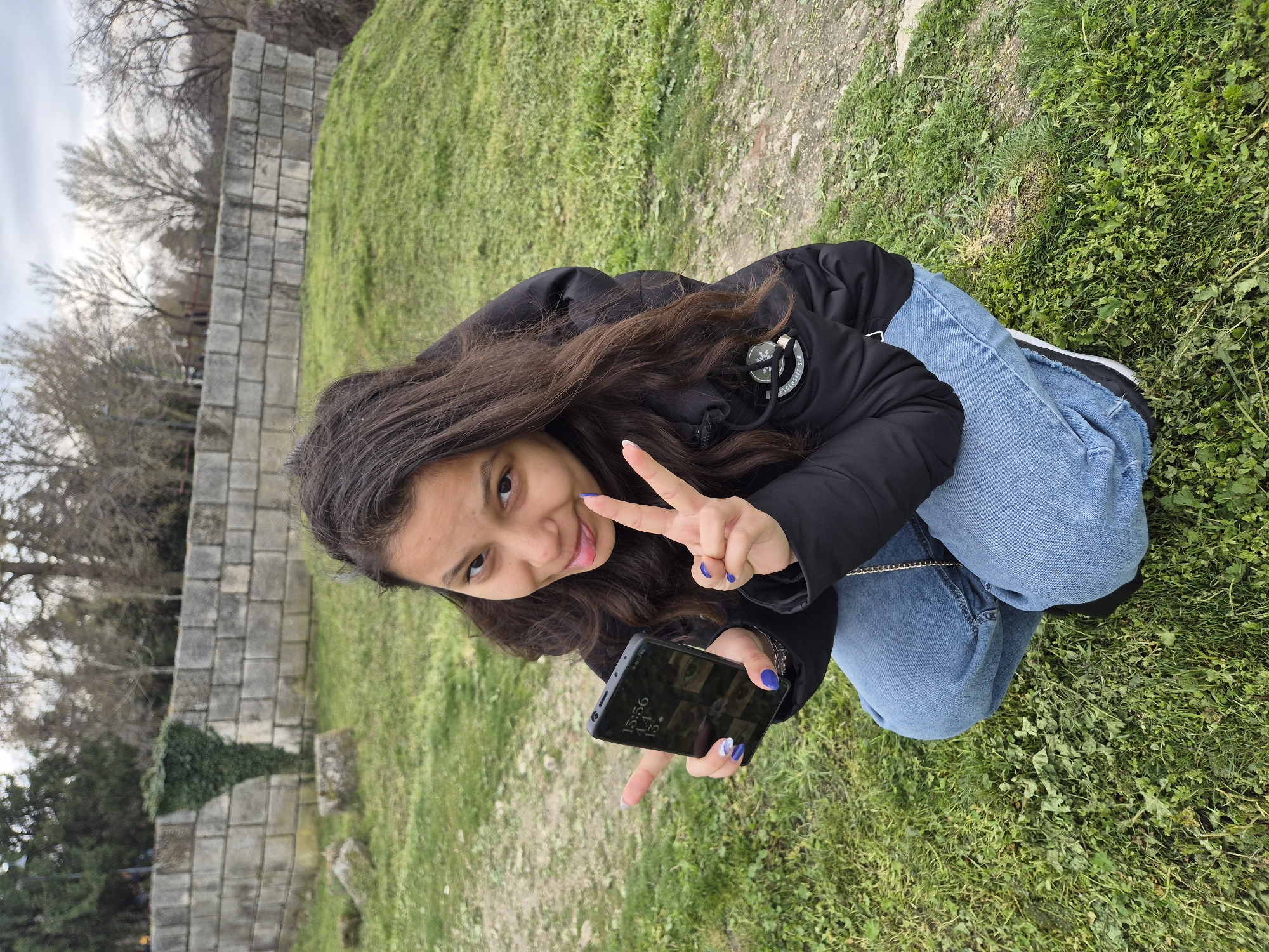 A young woman is kneeling on a grassy area next to a low stone wall, presumably in Silistra, Bulgaria. She is holding a black smartphone in her left hand, with the screen displaying a time of 15:56, date 4.11, and a number 13. The woman is flashing a peace sign with her right hand, sticking her tongue out, and smiling, while her fingernails are painted a dark blue. She is wearing a black jacket and blue jeans. The location has trees, grass, and a wall.