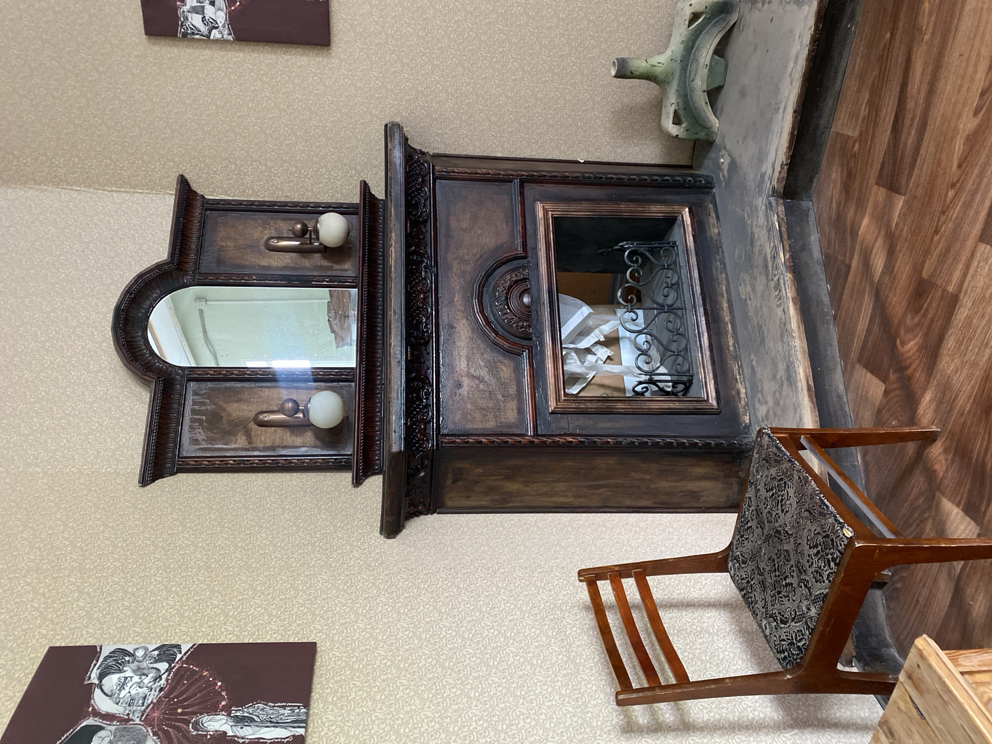 The image presents a partially rotated view of an interior setting. A dark, ornate wooden cabinet with a mirror and small cubby is positioned against a patterned beige wall. Above the cabinet are two framed artworks or prints. Adjacent to the cabinet and partially obscuring the wall is a wooden chair with a patterned cushion. A green and grey ceramic object sits atop the cabinet structure. The environment appears to be an indoor space. There is no visible human presence. The location is Yekaterinburg, Russia.