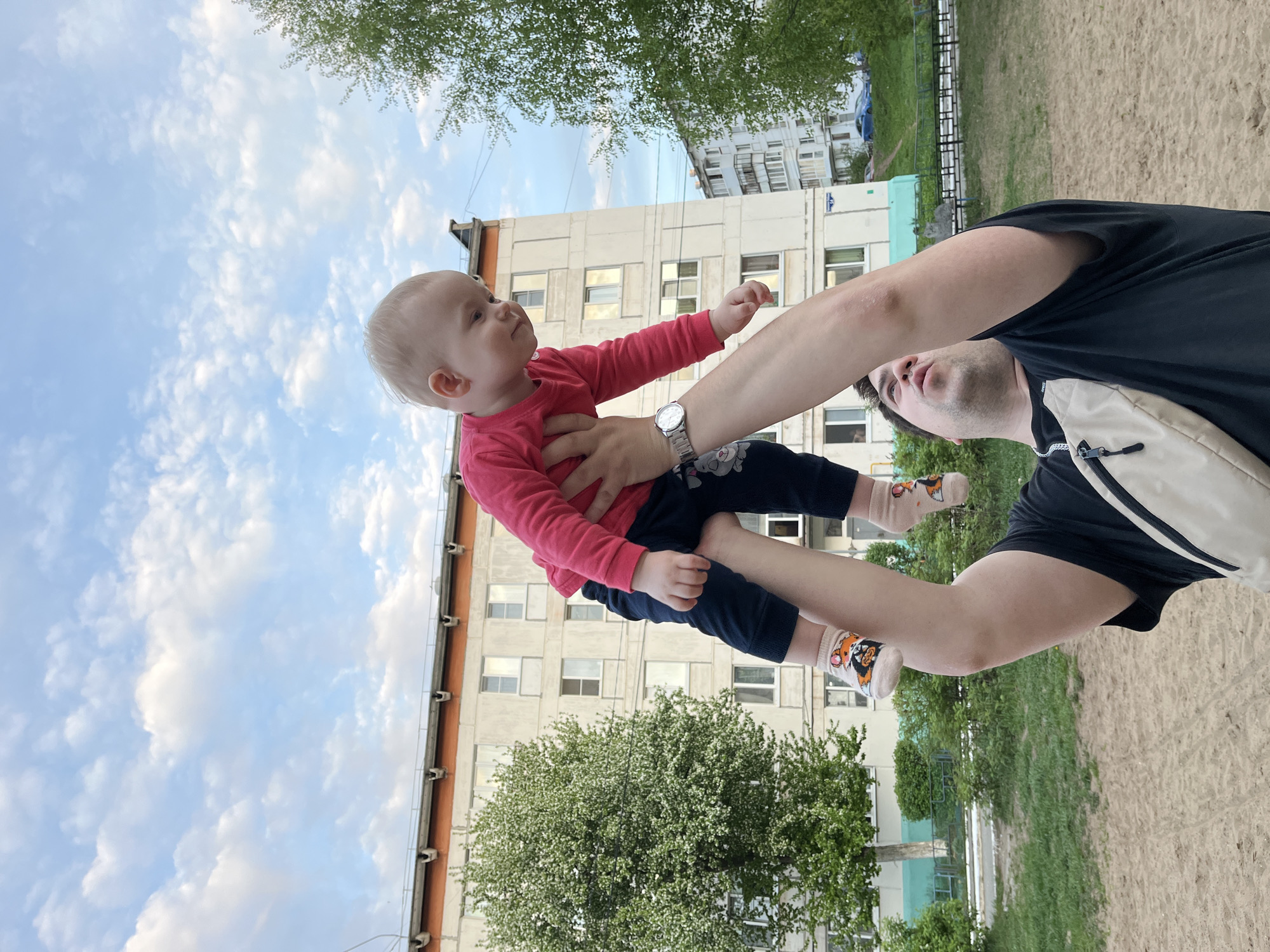 A man is holding a baby up in the air. The baby is wearing a pink shirt, dark blue pants, and socks with animal images. The man is wearing a black shirt, light-colored bag, and a watch. They are outdoors in a park or playground, with a sandy ground. A building is visible in the background. The sky is blue with clouds. The location is most likely in the city of Chaikovsky, Russia based on the building architecture and common urban landscape elements.