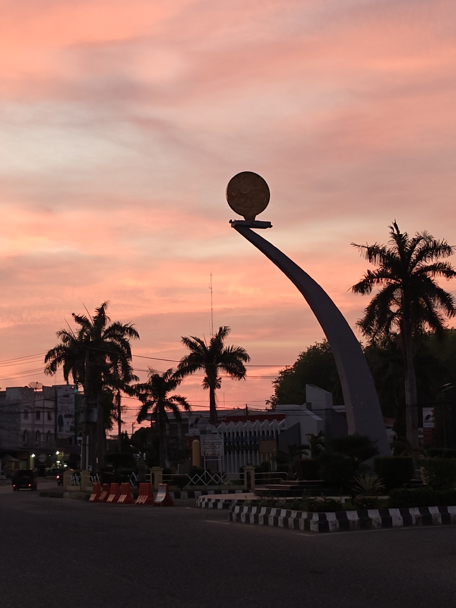 A long-shot captures a city roundabout in Lhokseumawe, Indonesia at dawn or dusk. The sky is a blend of light pink and peach hues. Dominating the scene is a tall, gray monument with a large circular emblem at its apex. Silhouetted palm trees stand to the sides. A line of orange and white traffic barriers is positioned along the curb. Beyond the roundabout, buildings are visible. The road curves around the roundabout. A vehicle can be seen on the road.