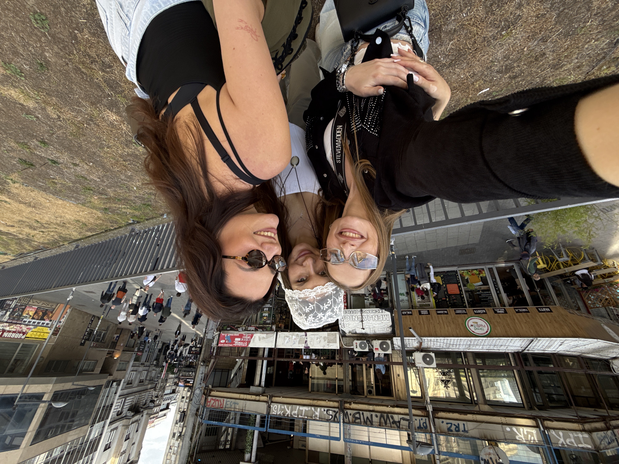 Three women are in a selfie, with an upside-down background of a city street. The woman on the left has brown hair and is wearing sunglasses and a black top. The woman in the center has on a white lace hat and is wearing a silver necklace. The woman on the right has blonde hair and is wearing glasses and a black cardigan. In the background is a street scene with shops, buildings, and people walking. The image is from Belgrade, Serbia, indicated by the signage in the background (e.g., "Menjačnica Exchange Office"). A restaurant named "Street Pasta Bar" with "ROSE HIP..." on the sign is visible. There is graffiti on the buildings.