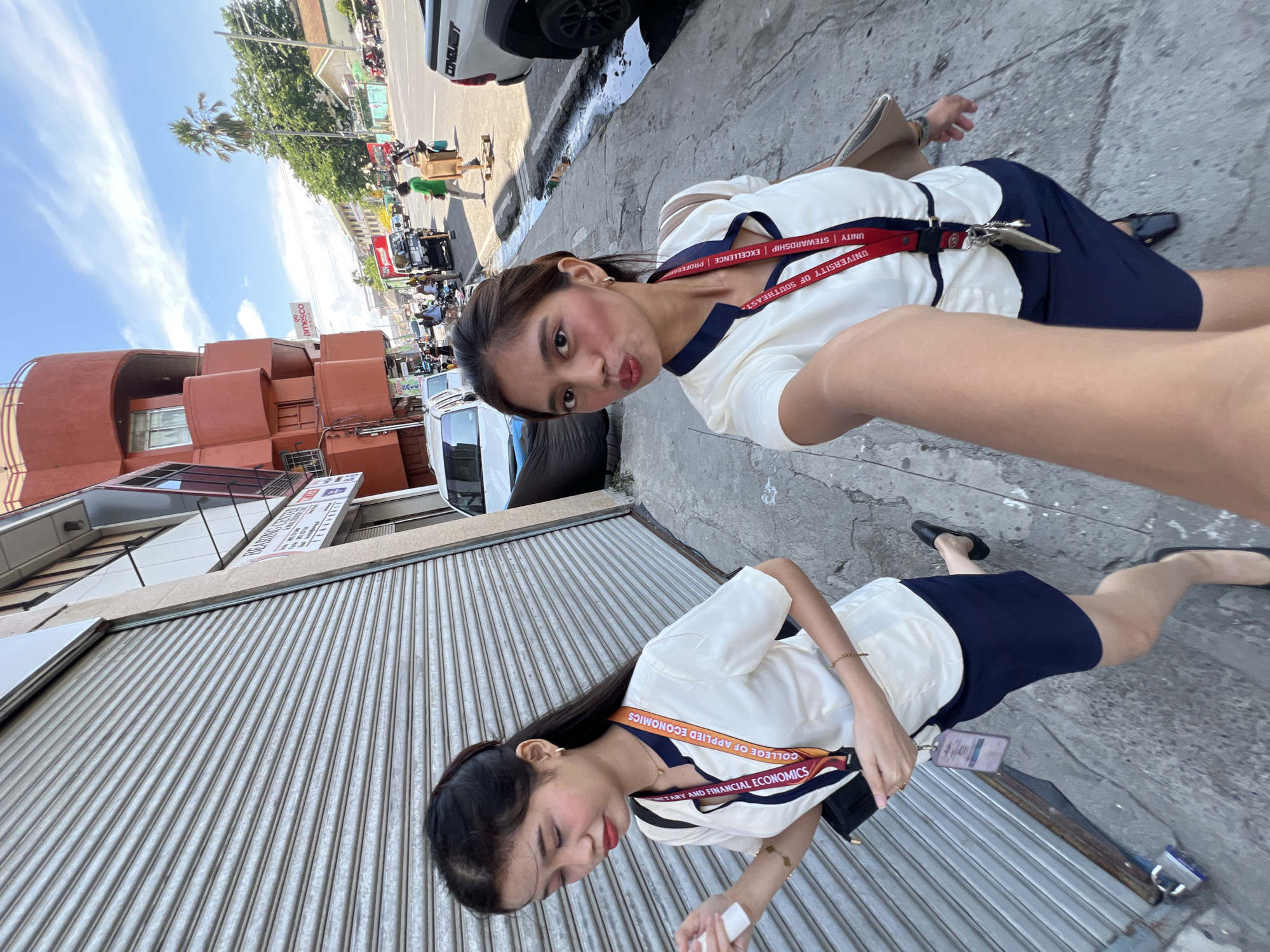 In a medium-shot, selfie-style photograph, two young women stand on a sidewalk in what appears to be a commercial district of Davao City, Philippines. The woman in the foreground, facing the camera, is making a kissing gesture. Both women are wearing matching white polo shirts with navy collars and navy skirts. They also have red lanyards around their necks with university insignia visible. The woman on the left is looking down and away from the camera, while the woman on the right is looking directly at the camera.
Behind them, a gray metal roll-up door covers the storefront of a building. The building itself has an orange, curved architectural design on the upper floors. Beyond the immediate storefront, a street is visible with several vehicles, including cars and motorcycles, and pedestrians. Trees and other buildings line the street, indicating an urban environment. The sky is blue with scattered clouds, suggesting a daytime scene. The sidewalk appears to be made of concrete and shows some wear and tear, with a gutter visible along the edge of the street. The overall impression is of a casual, everyday scene in a bustling city.