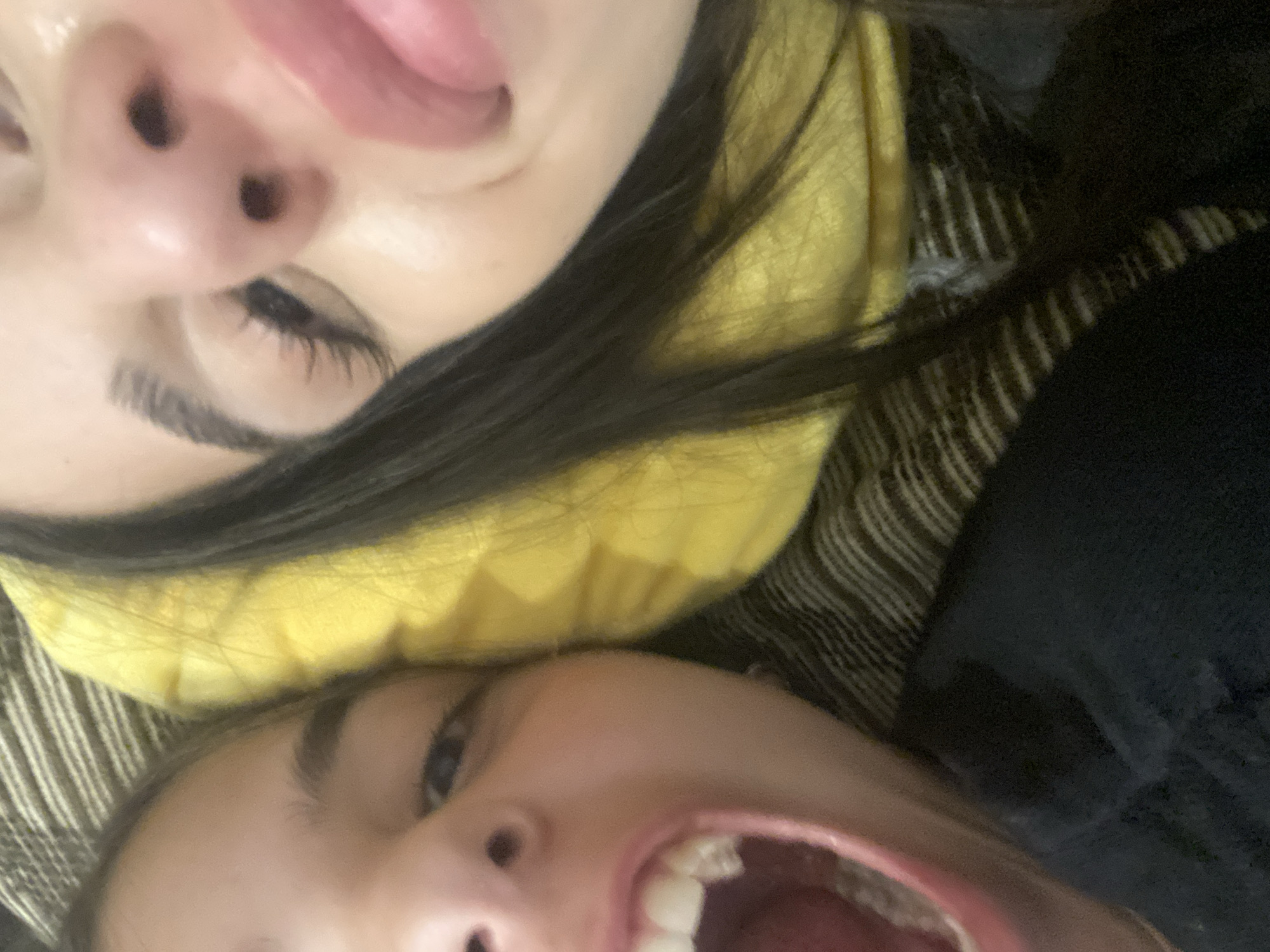 Two individuals are captured in a close-up, unconventional composition. The upper portion of the image shows the face of an individual with a visible nose, eye, and tongue protruding from their mouth. The individual has dark hair and is resting on a yellow object. Beneath this individual is another person with their mouth open, revealing their teeth. The picture is taken at an extreme angle. No information indicates any location in Dragovishtitsa, Bulgaria.