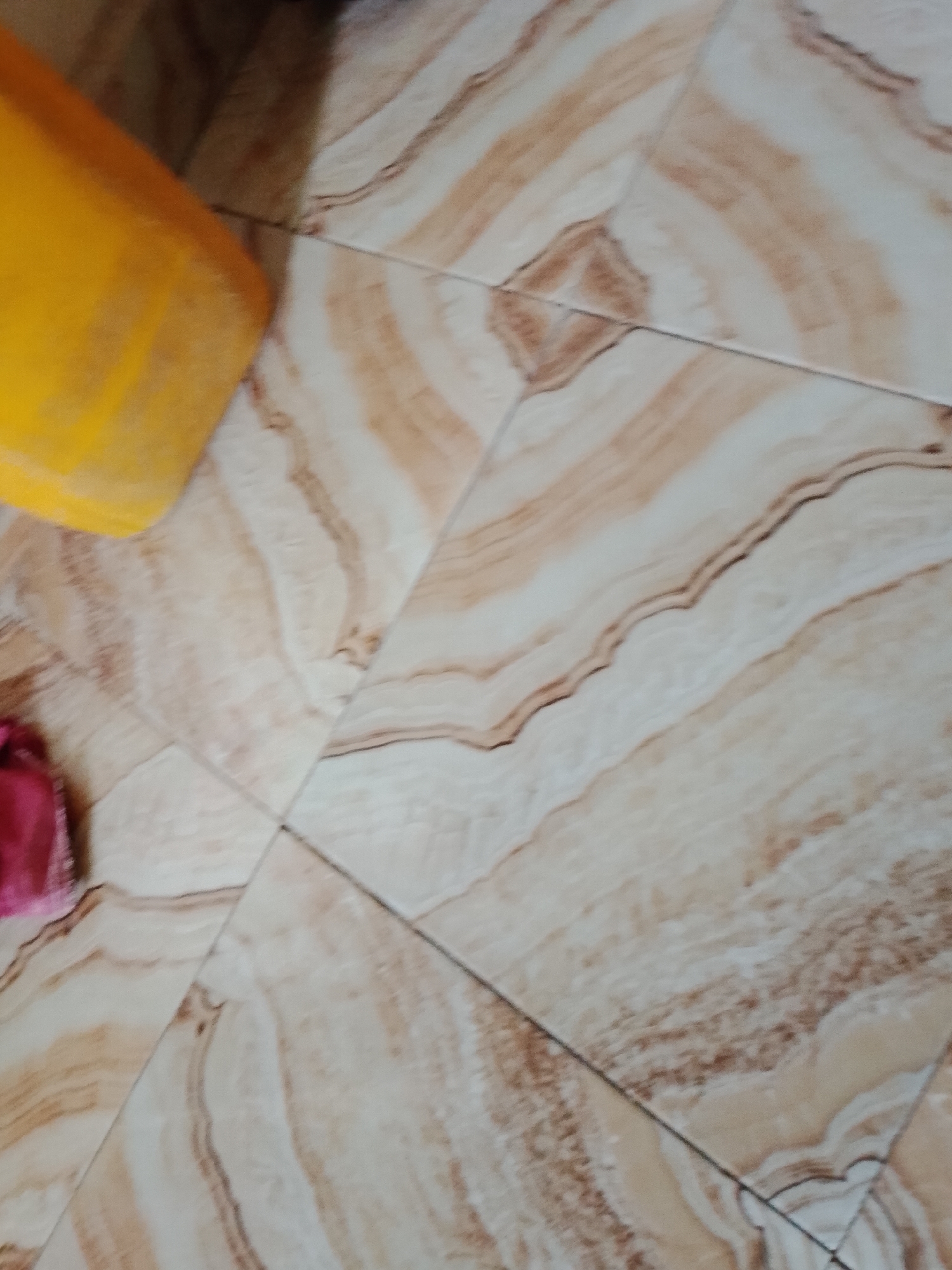 The image shows a close-up, angled view of a tiled floor. The tiles have a marble-like pattern with beige, cream, and light brown veining. In the upper left quadrant, a portion of a bright yellow bucket or container is visible. To the left of the bucket, a small section of a pink or red fabric, possibly a piece of clothing or a cloth, is present. The scene is outdoors or in a well-lit indoor space with natural light. There is no visible indication of the city of Pantinapu, Nigeria, or any specific landmarks.