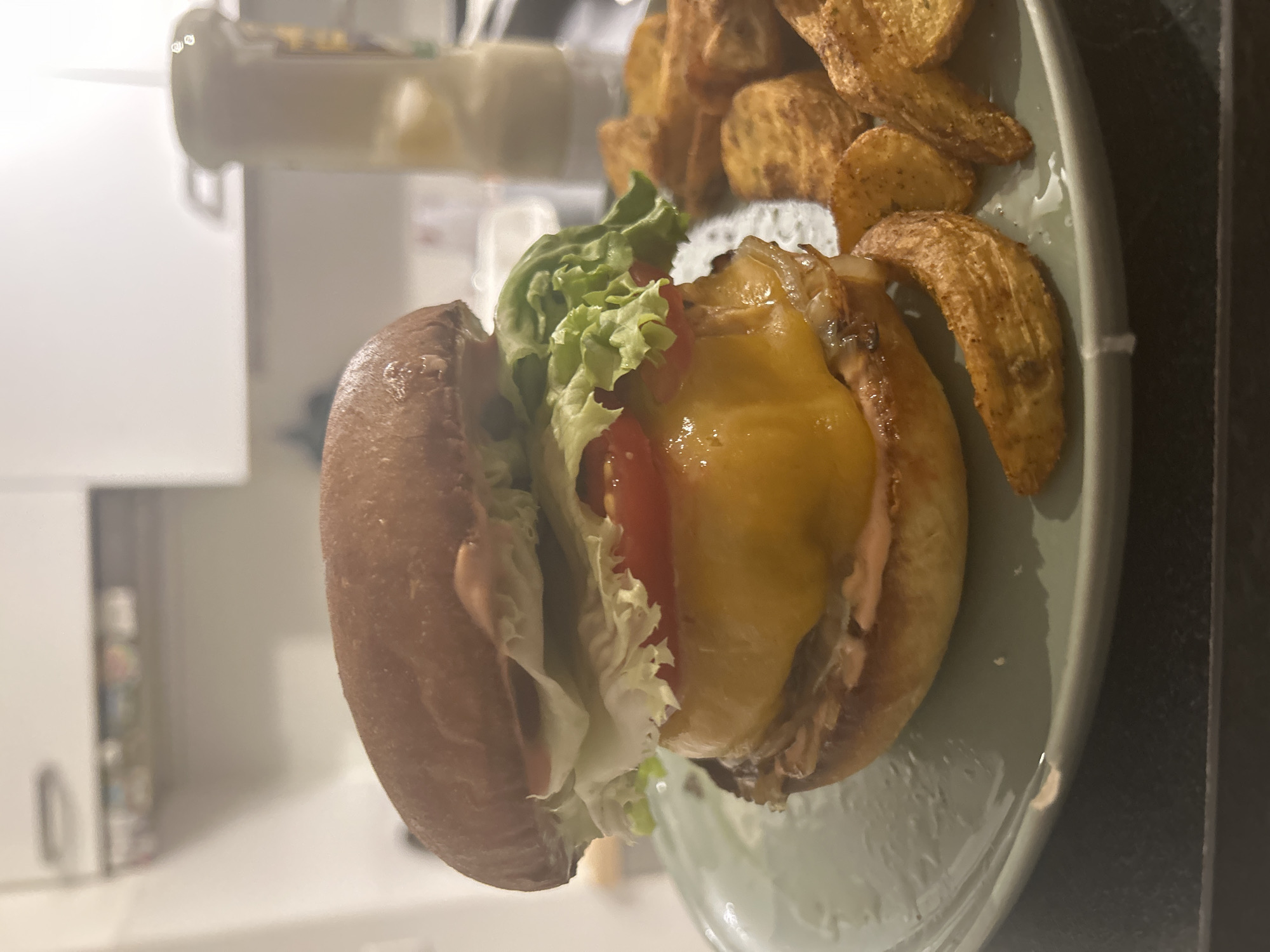 A burger with lettuce, tomato, cheese, and a pink sauce, sits on a light green plate next to potato wedges. The burger is in the center of the plate and positioned towards the left edge. Potato wedges fill the right side of the plate, with a mix of sizes and shapes. The plate is partially set on a dark surface in front of a white kitchen cabinet. The location is not discernible from the image.