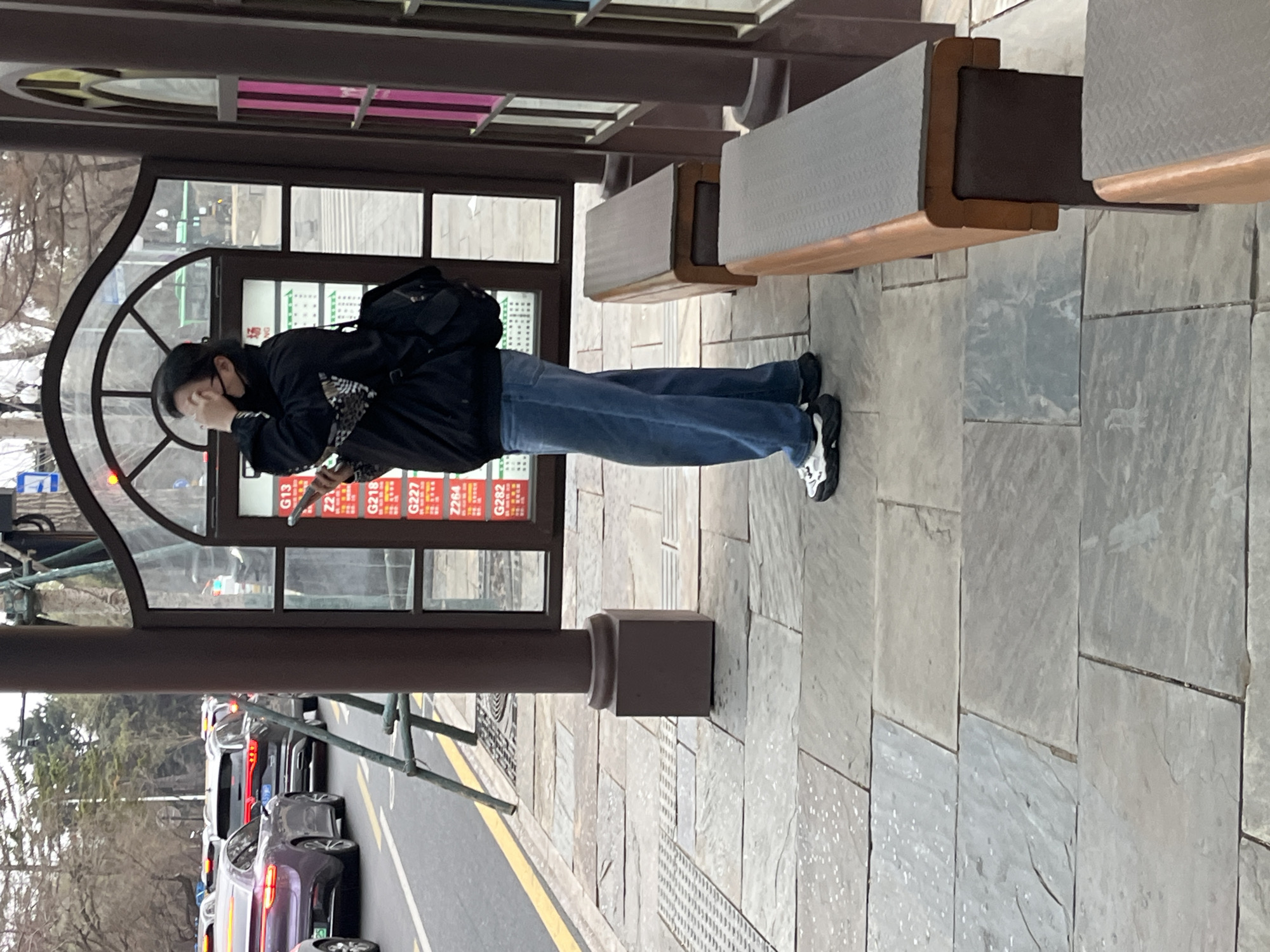 The image shows a bus stop in Changchun City, China. A person wearing a black jacket, blue jeans, and black and white sneakers is standing inside the bus stop, looking at their phone. They are wearing a black face mask. The bus stop has seats made of light brown wood and light gray metal. A street with cars is visible outside the bus stop. The day appears overcast. Bus route information is displayed on the glass inside the bus stop.