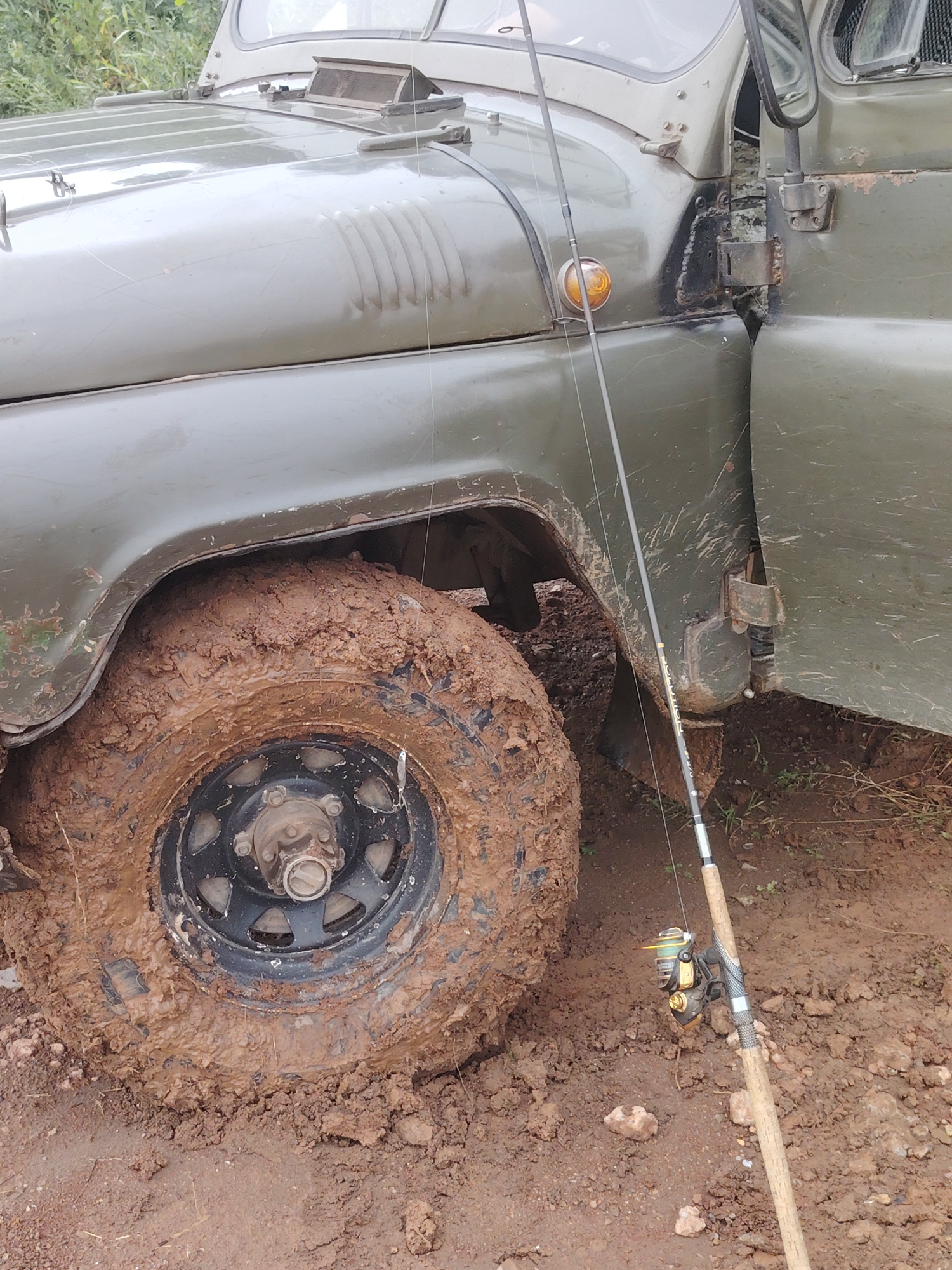 The image depicts a muddy off-road vehicle, likely a UAZ, parked on a dirt track in what appears to be a rural or wilderness area. The vehicle is predominantly green, with visible mud splatters on the tire, fender, and door. The front passenger side tire is heavily caked in mud. A fishing rod with a reel is propped up against the vehicle, leaning against the door. The ground is a mixture of mud, dirt, and some sparse vegetation. The background shows a hint of greenery, suggesting an outdoor environment. The lighting is overcast, creating a muted color palette. No people are present in the image. The location is identified as Прикамский, Russia, although this specific detail cannot be independently verified from the visual information alone.