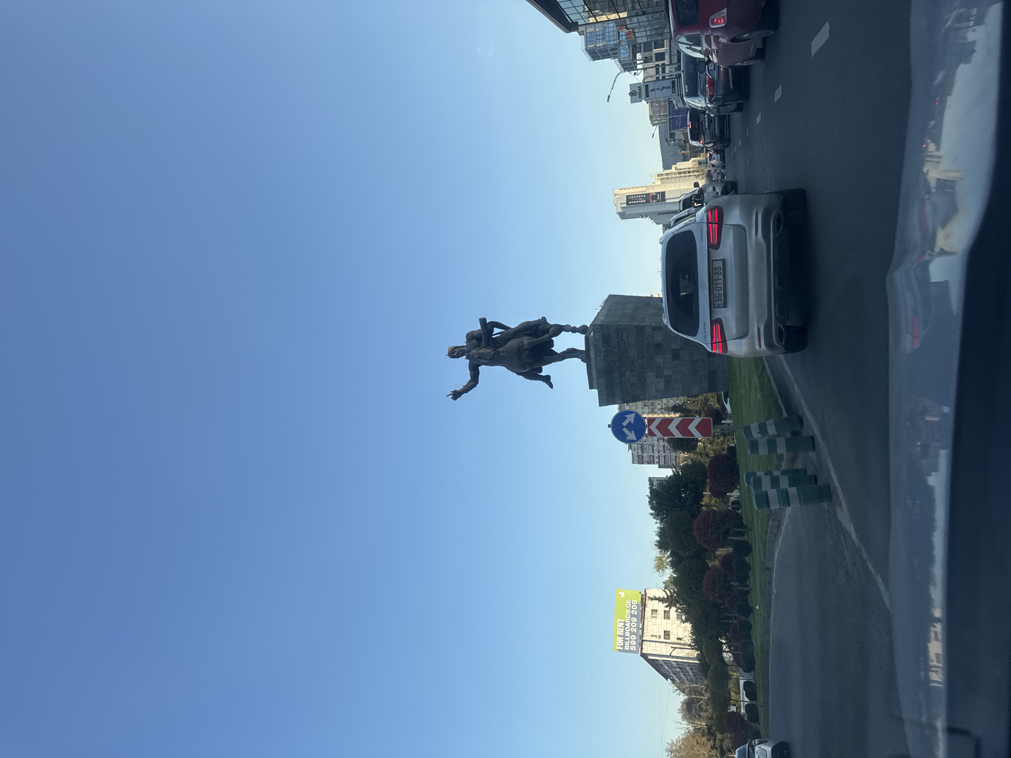 The image captures an urban street scene from the perspective of a vehicle, looking towards a prominent equestrian monument under a clear blue sky. The monument, positioned on a stone pedestal, features a large bronze statue of a man on horseback, holding a staff or cross, with another figure appearing below the horse.
To the right of the monument, a multi-lane road is busy with traffic. In the foreground, a silver SUV, possibly a Porsche Macan, displays a Georgian license plate "BG-011-BB." Further down the road, additional cars are visible in traffic. Road infrastructure includes a paved surface with lane markings, and a median strip adorned with green and white striped bollards and manicured shrubs. A blue circular street sign with a white arrow and a red and white chevron sign are also present.
Tall, modern buildings with reflective facades line both sides of the street in the background, characteristic of a city environment. On one building, a brightly colored billboard advertises "FOR RENT BILLBOARDS.GE" with the repeated phone number "599 209 209".
The scene is bathed in bright daylight, indicating a sunny day, likely late morning or early afternoon, with no clouds in the sky. The overall atmosphere suggests a typical, bustling day in a city identified as Tbilisi, Georgia. No specific emotional content is discernible from the image.