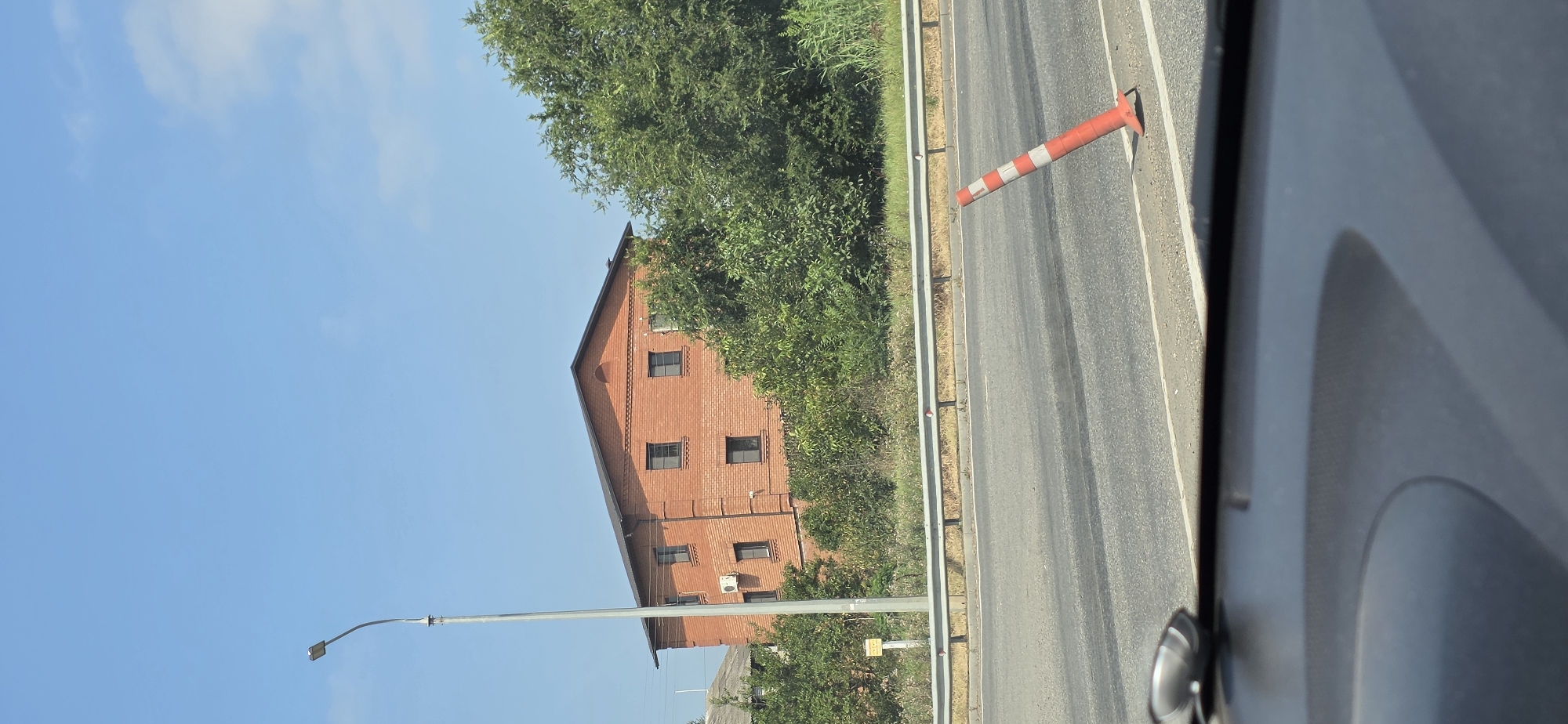 A bright, clear day in Mineralnye Vody, Russia, as seen from inside a vehicle. The foreground is dominated by the car's dashboard and windshield reflection. Beyond that, a two-lane asphalt road stretches out, marked by white lines. A bright orange and white traffic cone lies tilted on the right side of the road. A metal guardrail separates the road from a grassy embankment dotted with various trees and shrubbery. Behind the embankment, a three-story red brick building with a dark roof and evenly spaced windows stands under a clear blue sky. A tall, grey lamppost is visible to the left of the building.