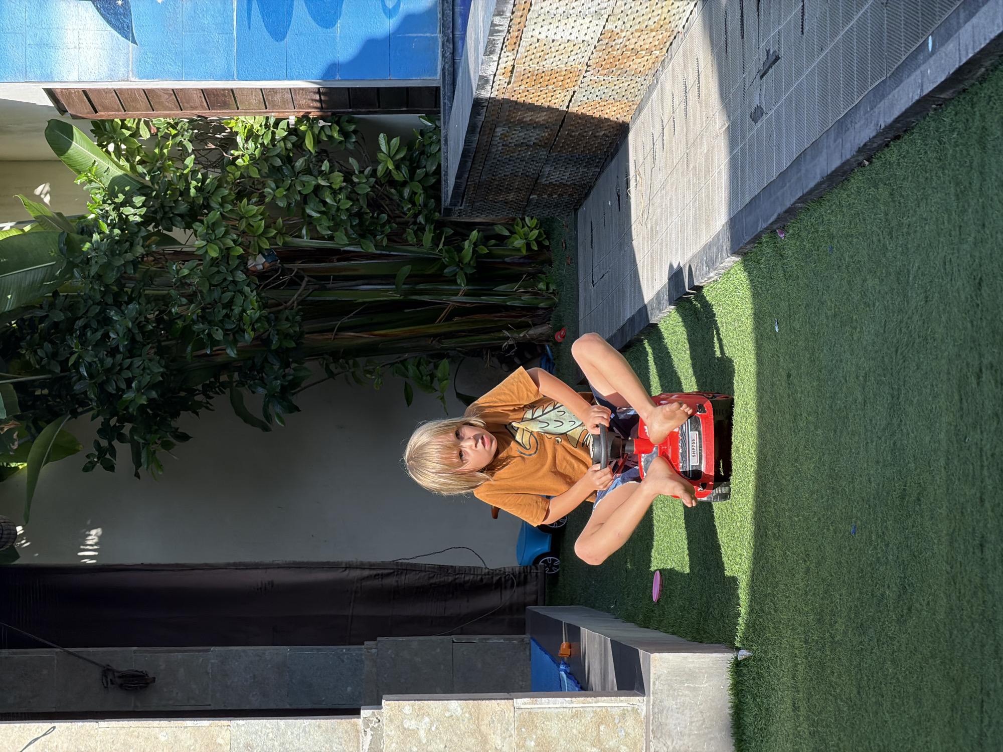 A young child with blonde hair sits on a red toy car on a patch of artificial grass. The child wears an orange t-shirt and blue shorts. The setting appears to be an outdoor area adjacent to a building with a blue wall and various plants. There is a tiled surface and steps leading down to the level where the child is located. The scene suggests a backyard or courtyard setting, possibly in Kuta, Indonesia.