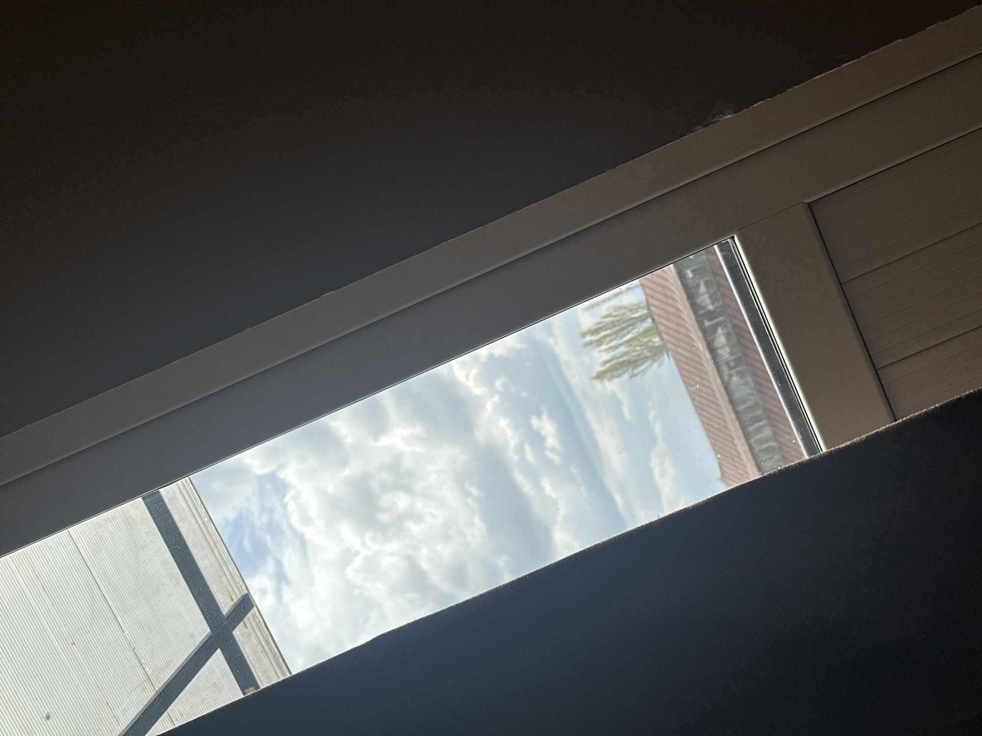 A view through a rectangular window shows a sky with puffy white clouds, partially obscured by buildings. On the left, a light-colored structure with black cross-bracing is visible. On the right, the side of a building with a brick-like pattern can be seen. The interior framing the window consists of a white frame, dark gray walls, and ceiling. The scene's location is Unknown, Unknown.