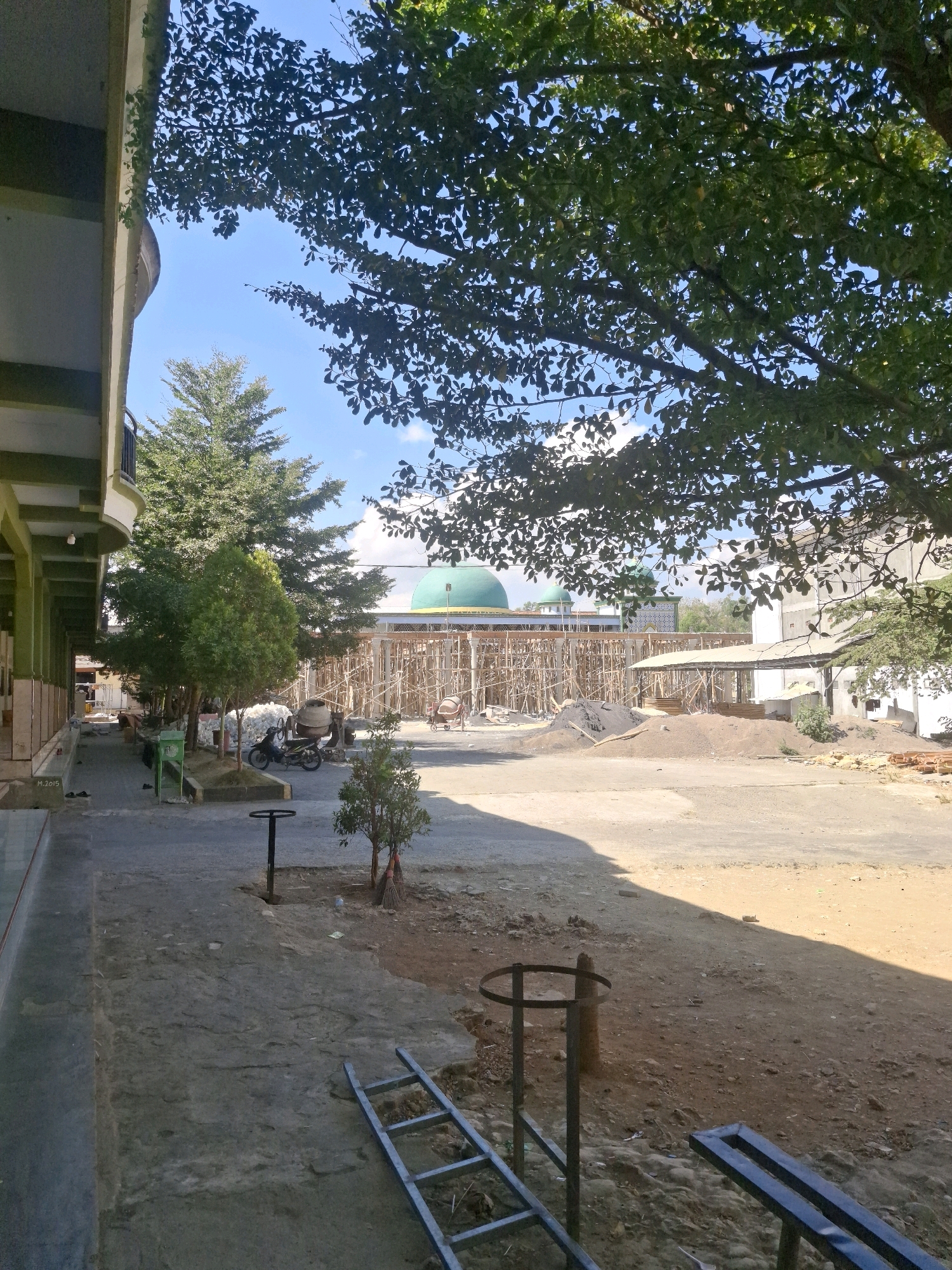This is a photograph taken outdoors in Potoan Laok, Indonesia, depicting a construction site in front of a building with a green dome, likely a mosque. The foreground features a paved walkway running alongside a building with columns and a partial view of a concrete structure. To the right of the walkway, the ground is unpaved and sandy, with piles of construction materials, including sand and what appears to be a cement mixer. A motorcycle is parked on the paved area. Further back, scaffolding surrounds the construction of a building. Trees are present throughout the scene, with dense foliage from a large tree in the upper right casting shadows. The sky is blue with scattered clouds. There are no people visible in the scene.