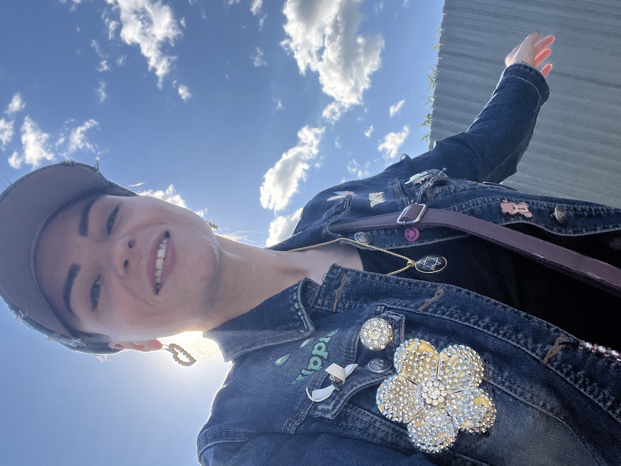 In Krasnoe-na-Volge, Russia, a woman wearing a denim jacket and a cap poses for a selfie outdoors against a bright blue sky with scattered clouds. The woman has her hair pulled back and is smiling. She is wearing a necklace with a Star of David pendant and several pins on her jacket, including a large flower-shaped brooch adorned with rhinestones, a pin with a white ribbon and a small brown object, and other decorative pins. Her jacket has the word "hippie" embroidered in green on the collar. A purple strap from a bag is visible across her chest. The background shows a grey, corrugated metal fence and some green foliage. The sun is bright, creating a lens flare effect.