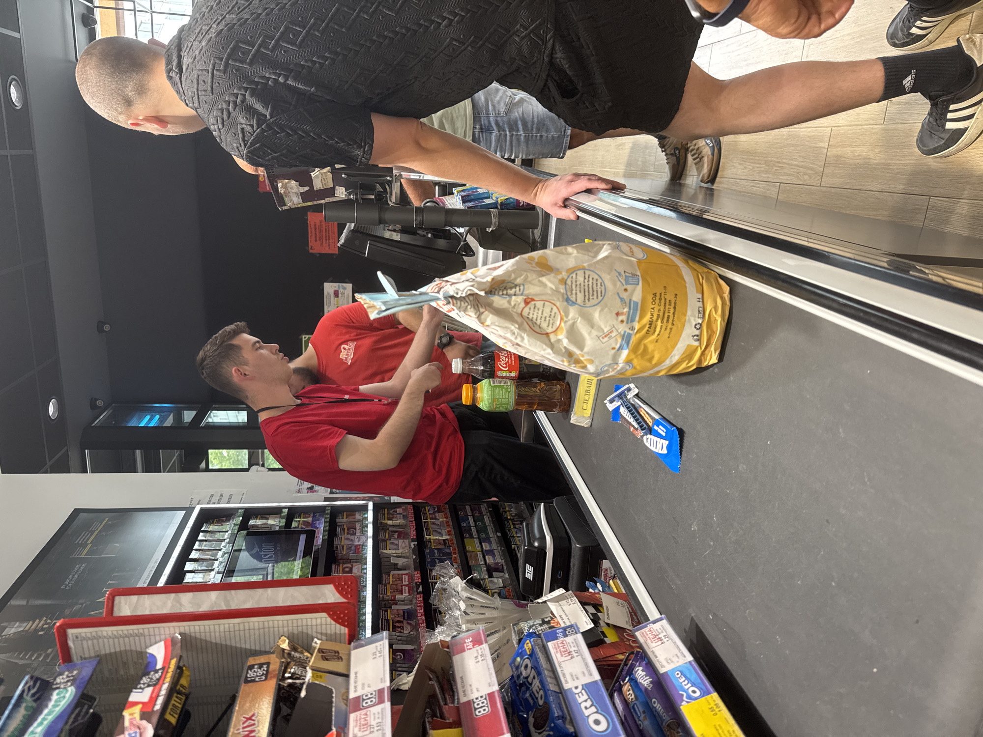 In Varna, Bulgaria, inside a brightly lit convenience store with tiled flooring and a black suspended ceiling, a cashier in a red shirt is scanning items on a conveyor belt. To the right of the cashier, a customer wearing a black patterned shirt and black shorts is waiting for their items to be processed. A large bag of goods is positioned prominently on the conveyor belt, with a bottle of Coca-Cola and a bottle of iced tea beside it. Various snack items, including Oreo cookies and Milka chocolate bars, are visible on shelves to the left of the scene. The overall environment suggests a typical retail transaction at a point of sale.
