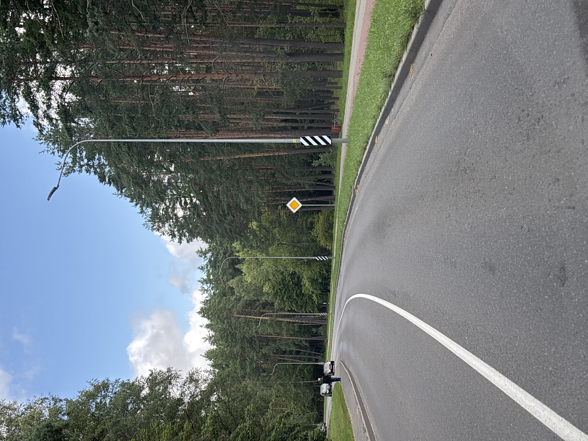The image depicts a paved road in a wooded area, possibly on the outskirts of Ждановіцкі сельскі Савет, Belarus. The road curves to the left and is lined with a white dashed line on the right side. On the left side of the road, there is a sidewalk or pedestrian path. Two cars are parked on the left side of the road, and a person is walking on the sidewalk.
The environment is characterized by a dense forest of tall pine trees bordering the road on the right. On the left, there are also trees, but they appear less dense. A street lamp is visible on the right side of the road, and another is further in the distance. A diamond-shaped yellow traffic sign with a black border is posted on the right side of the road, as is a striped pole with the number "12" on it. The sky is blue with scattered white clouds. The ground is covered with grass along the sides of the road. The overall scene suggests a semi-rural or park-like setting within or near the village of Ждановіцкі сельскі Савет.