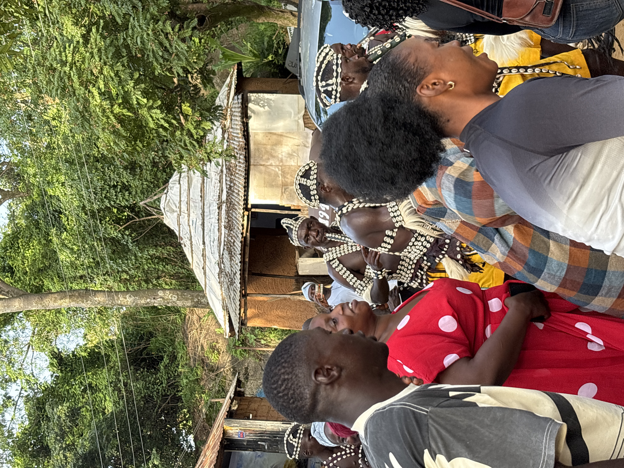 This image depicts a group of people, some adorned in traditional attire featuring shell necklaces and headbands. The individuals are gathered outdoors, with lush green trees and structures made of corrugated metal and wood suggesting a rural or semi-rural setting. The time of day appears to be late afternoon or early evening, given the warm, diffused lighting.
The people are looking in various directions, with some engaged in conversation or observing something beyond the frame. One woman in the foreground wears a red dress with white polka dots, while another person is seen from behind wearing a black and white t-shirt. The overall mood seems to be one of community and perhaps a cultural gathering or celebration.
Visible text in the image includes numbers like "200", "199", and "20", which are likely part of signage or markings on objects within the scene, although their specific meaning is unclear without further context. The presence of traditional clothing, coupled with the natural surroundings and community interaction, points to a scene rooted in local culture.