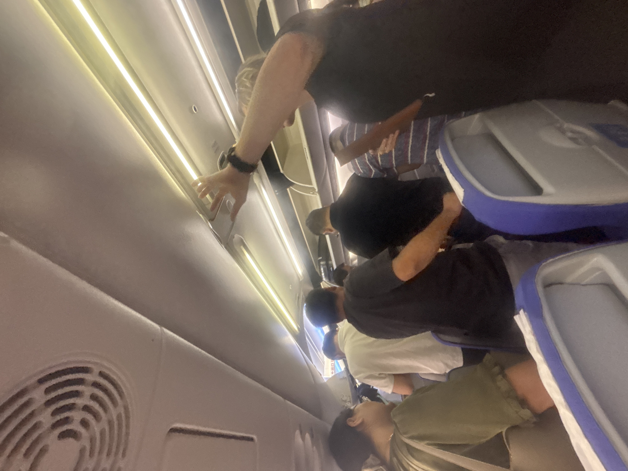 The image shows a group of people standing in the aisle of an airplane, likely boarding or disembarking. A woman in the foreground on the right, wearing a black top and a watch, is reaching up towards the overhead compartment. Other passengers are visible in the background, some standing and some seated in blue and white airline seats. The interior of the plane is lit by overhead fluorescent lights. There is a circular air vent visible on the left side of the ceiling. The image is taken from a low angle, looking upwards.
