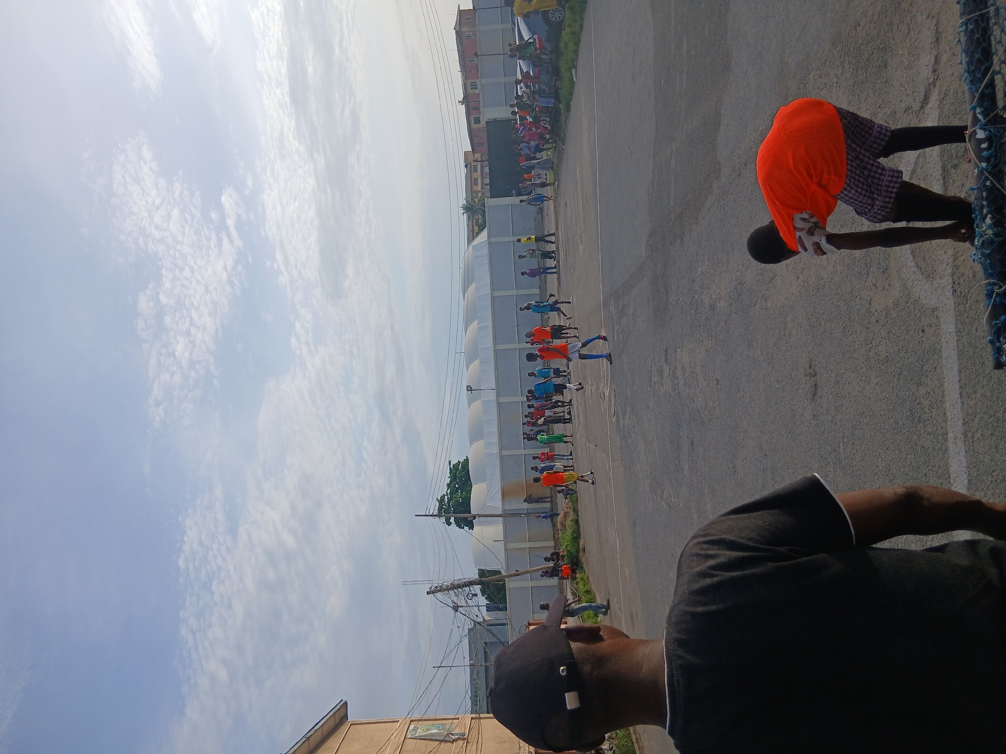 A medium-angle, outdoor shot shows a street scene, presumably in Lagos, Nigeria, with a group of people preparing for some kind of race or activity, viewed from the perspective of someone standing near the edge of the road.
In the midground, a line of individuals is arranged along the side of the road, some wearing orange, blue, and other colored tops, suggesting some kind of team or event. In the right foreground is a man wearing an orange shirt and plaid shorts leaning over the edge of the road, possibly setting up equipment. To his left is a blue net. Closer to the lower right corner, the back of another man wearing a black shirt and baseball cap is visible, placing the viewer within the scene.
The background contains buildings and houses, possibly indicating an urban or suburban area. A large, curved white structure stretches along the road, possibly a greenhouse or warehouse, adding a unique element to the setting. Power lines are visible above. The sky is partly cloudy. The surface of the road appears to be asphalt.