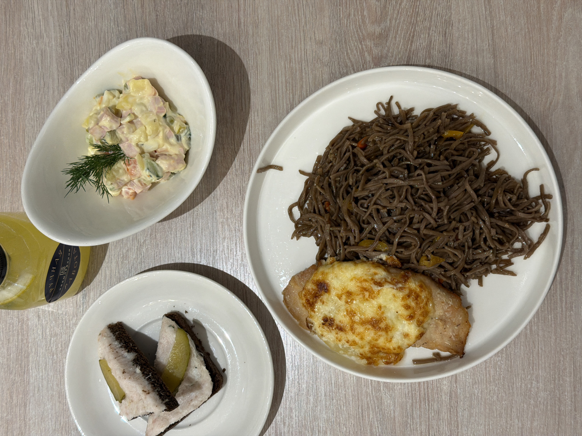 The image depicts a meal presented on a light-colored, possibly wooden, surface, likely a table. The meal consists of three plates and a bottle.
The plate at the top left contains a salad, potentially Russian salad (Olivier salad), garnished with a sprig of dill. The plate on the bottom left features two pieces of dark bread topped with what appears to be salo (cured pork fat) and a slice of pickle. The large plate on the right has brown noodles and a piece of meat topped with melted cheese.
The bottle, partially visible on the left, has a label with Cyrillic text. The environment appears to be indoors, perhaps in a restaurant. Based on the cuisine and the Cyrillic text on the bottle, the meal is likely being consumed in Russia, with Moscow being a possible location due to its significance as a major city and a hub for diverse culinary experiences.