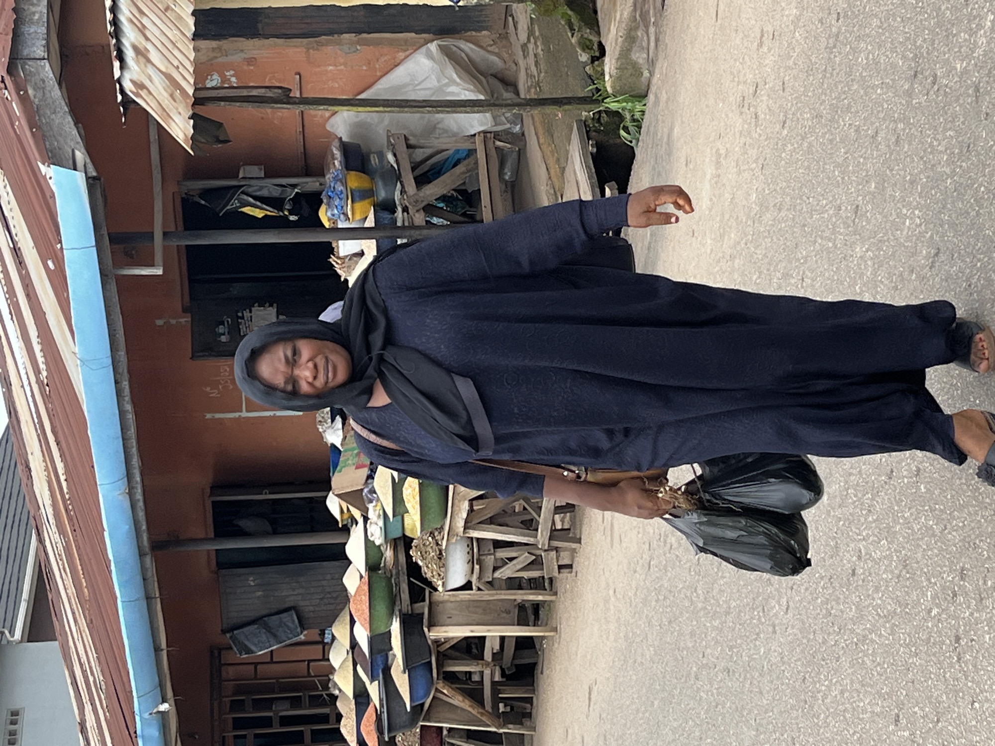 A woman is standing in a market, holding two black plastic bags in her right hand. She is wearing a dark blue abaya and a dark blue headscarf that covers her hair and neck. Her face is partially obscured by the headscarf, but her eyes are visible, and she appears to be looking towards the camera with a slight smile.
The market scene behind her shows stalls with various goods displayed. There are wooden structures, metal sheeting, and what appear to be colorful textiles or woven materials. Some items are stacked on shelves and in baskets, suggesting a variety of items for sale. The ground is paved, indicating an outdoor market. The light suggests it is daytime, possibly late morning or early afternoon, with no strong shadows indicating a bright sun. The overall impression is of a bustling local marketplace. The context provided is Jos, Nigeria. There is no visible text in the image.