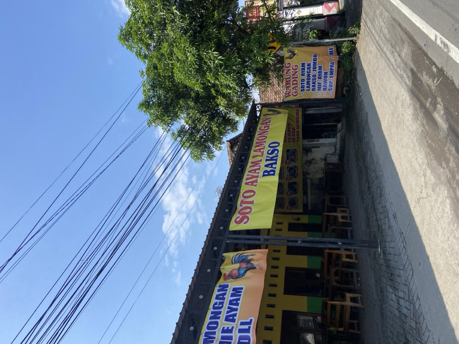 The scene shows a street-side eatery with a sign that reads "Soto Ayam Lamongan Bakso". The eatery has a roof and a yellow wall with a green trim. There are several empty tables with chairs in front of the eatery. A sign with the words "Warung Gading" lists various dishes. The image was taken in Pungging, Indonesia at latitude: -7.488, longitude: 112.561. There is a large tree with lush green foliage in the background. There is a blue sky with white clouds and several power lines running across the image. The sidewalk in front of the eatery is made of gray concrete blocks. The street in front of the sidewalk is paved with asphalt.