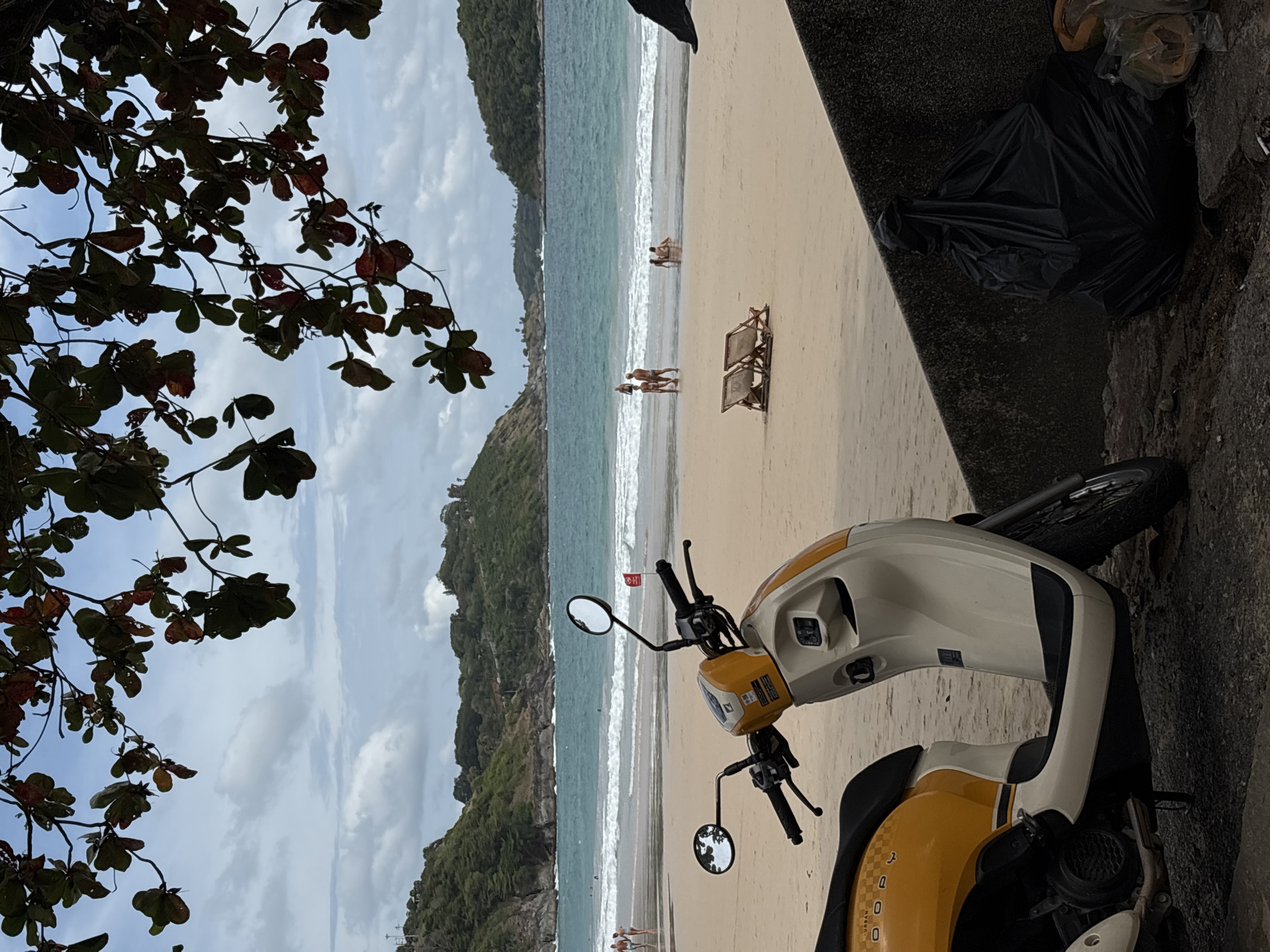 The image shows a view of a beach in Rawai, Thailand, framed by foliage on the left and a concrete wall and a parked scooter on the right. The beach features light sand and turquoise water with gentle waves. Several people are visible on the beach near the shoreline. In the foreground, a white and yellow scooter is parked beside a concrete wall, with a black plastic bag of garbage situated near it. The background includes verdant, hilly terrain.