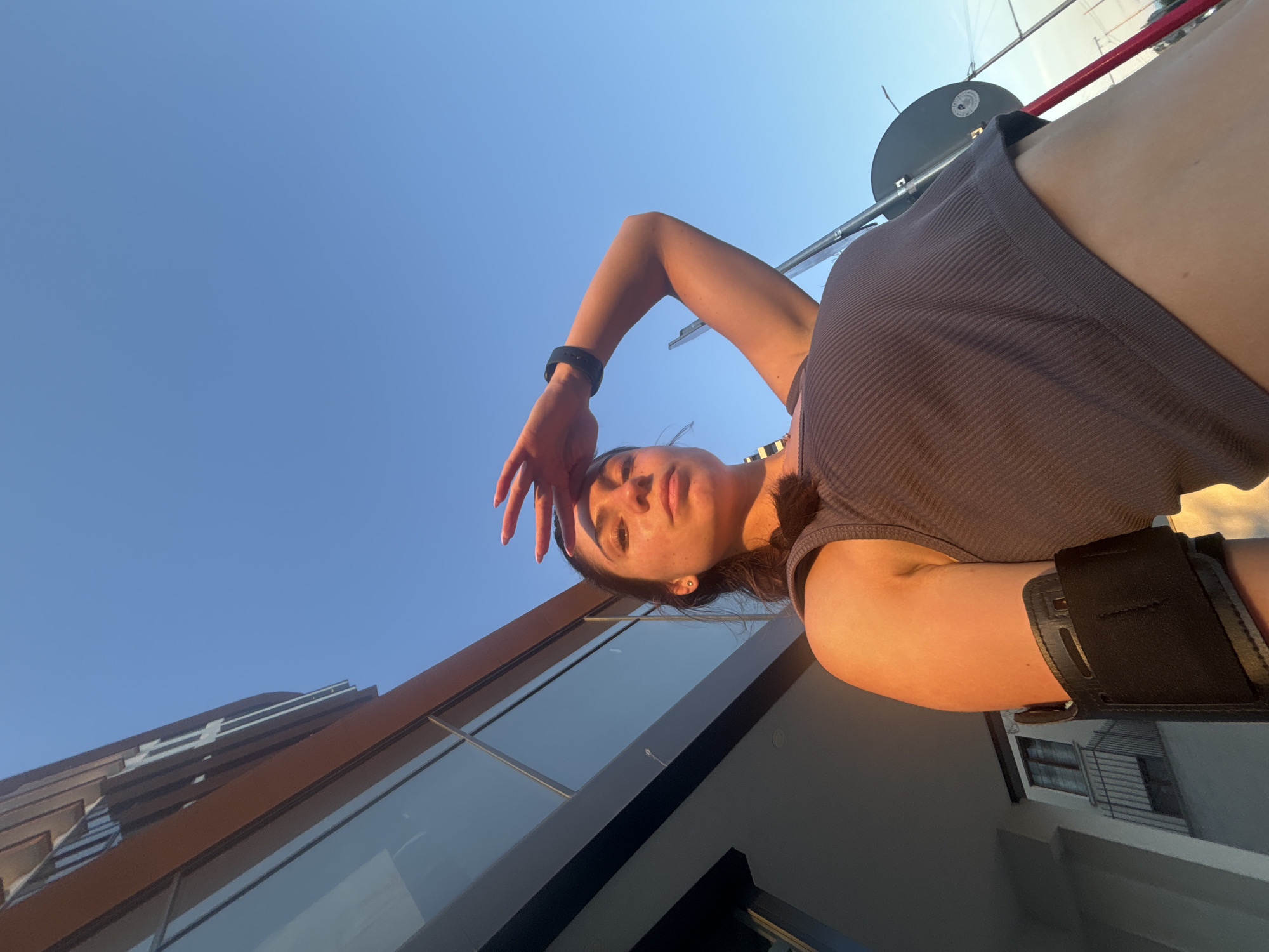A low-angle, full-shot photograph captures a young woman from the chest up, posed outdoors against a clear, bright blue sky. She stands in front of a modern building with a warm, brown-toned facade and large windows. The woman is wearing a dark grey ribbed tank top and has a black armband on her left bicep. Her right hand is raised to her forehead, shielding her eyes from the sun. Sunlight illuminates her face and right arm, creating a warm glow, while the left side of her body and the building are in shadow. A dark metal pole and part of a black circular object are visible to her right, likely part of an outdoor structure or fixture. The scene appears to be set in a contemporary urban environment, potentially a balcony or patio area of a building in Varna, Bulgaria.