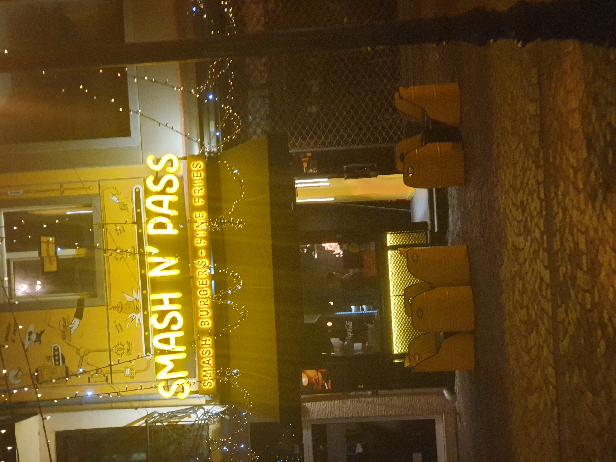 The image shows the exterior of "Smash N' Pass," a restaurant serving smash burgers and fine fries, located in Plovdiv, Bulgaria. The restaurant's bright yellow signage and decorations with strings of lights are prominent. The restaurant's facade includes windows showing interior scenes, including what appears to be staff and possibly customers inside. Stacked yellow chairs are visible outside.