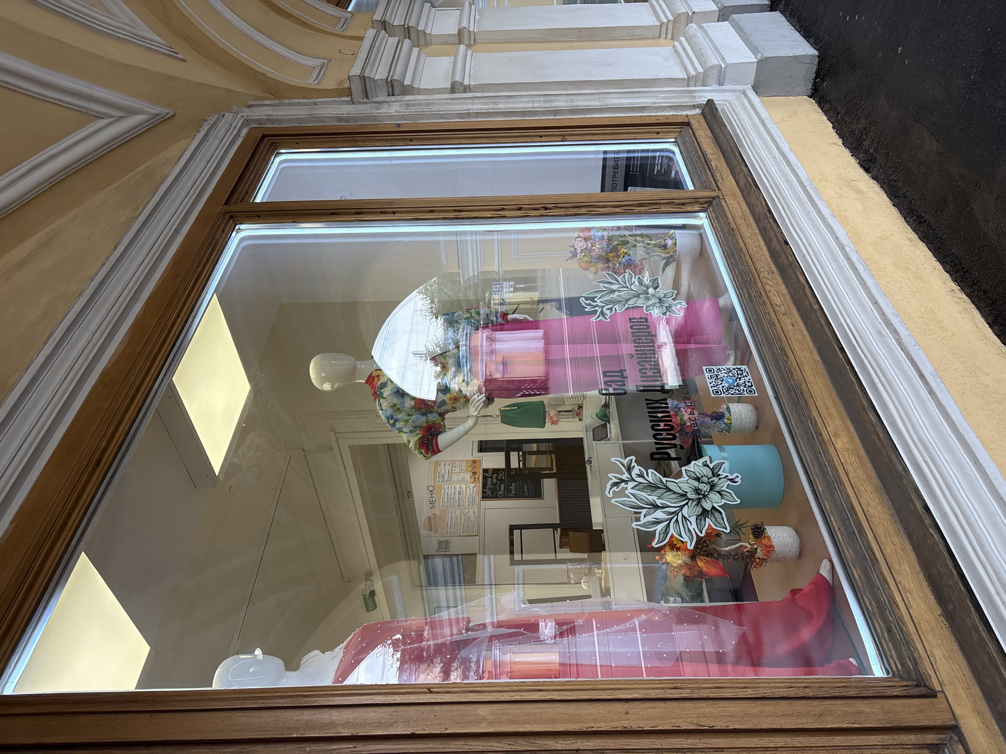 The image shows a shop window display in Saint Petersburg, Russia. The window frame is made of wood and the building exterior is painted in yellow and white. Inside the shop, there are mannequins dressed in clothing, a menu board, shelves, and decorative elements. The window display includes flowers and a sign in Russian that reads "Сад Русских Дизайнеров" (Garden of Russian Designers). The reflection in the window shows the building opposite the shop. The scene is taken from a slightly low angle, emphasizing the architectural details of the building.