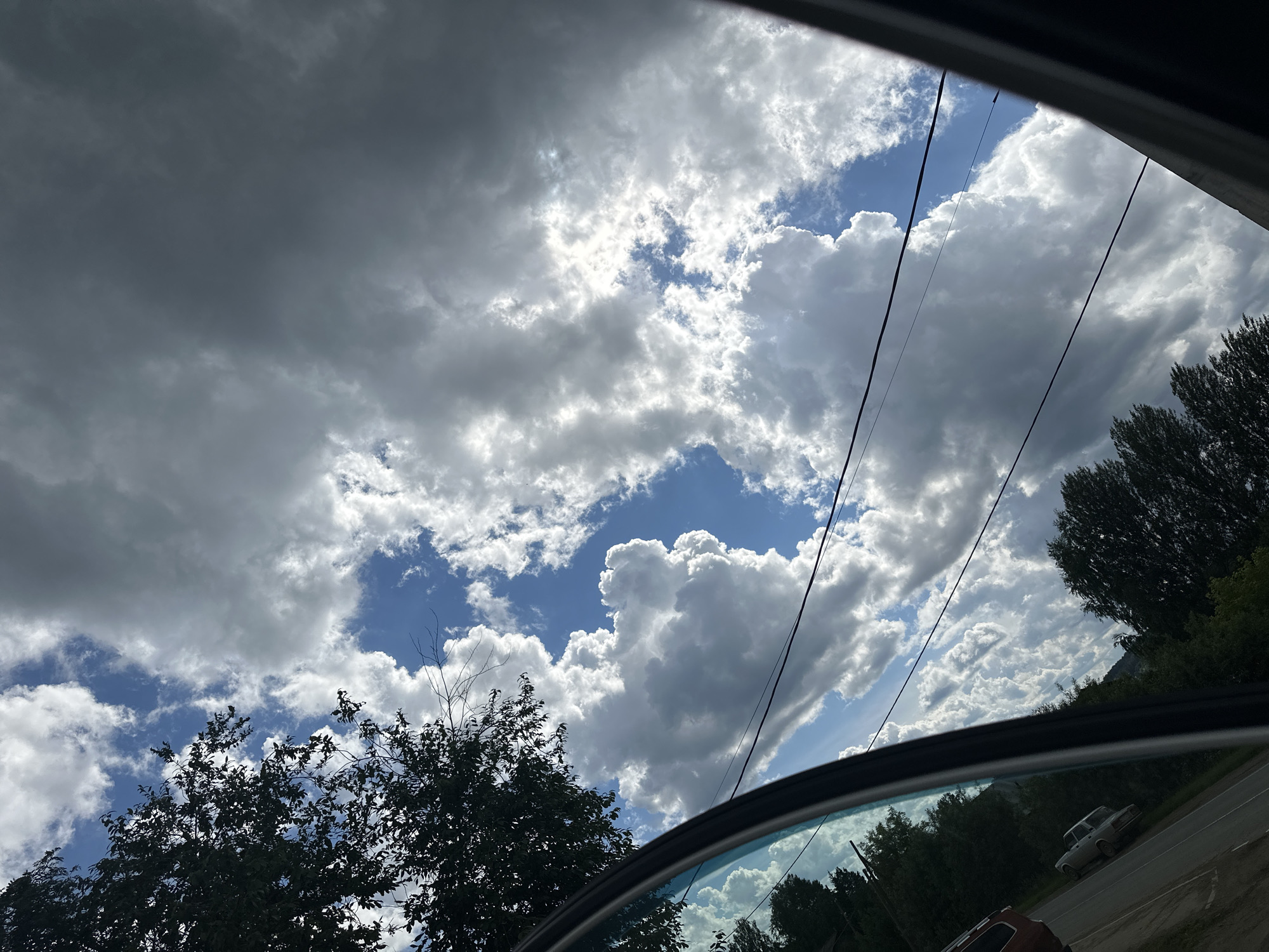 This is a landscape photograph taken from inside a vehicle, looking out through an open car door and window. The scene is dominated by a dramatic sky filled with a mixture of dark, heavy clouds and bright, fluffy white clouds. Patches of blue sky are visible between the clouds, with sunlight breaking through in some areas, suggesting a dynamic and possibly changing weather pattern.
In the foreground, the dark frame of the car door and window is visible, along with the curved reflection of the sky and trees on the car's window. To the left, the silhouette of dark green trees with dense foliage is prominent against the bright sky. To the right, more trees line a road, and a light-colored car is seen driving on this road. Power lines run diagonally across the frame, adding a man-made element to the natural scenery.
The overall impression is one of a journey in progress, with a view of a natural landscape under a complex sky. The lighting and cloud formations suggest it is daytime, possibly late morning or afternoon, with the sun intermittently obscured by clouds. The presence of trees, a road, and a car indicates a rural or suburban setting. There are no visible people or text in the image. The location context of Ilyinsky, Russia, implies the specific geographical setting for this scene.