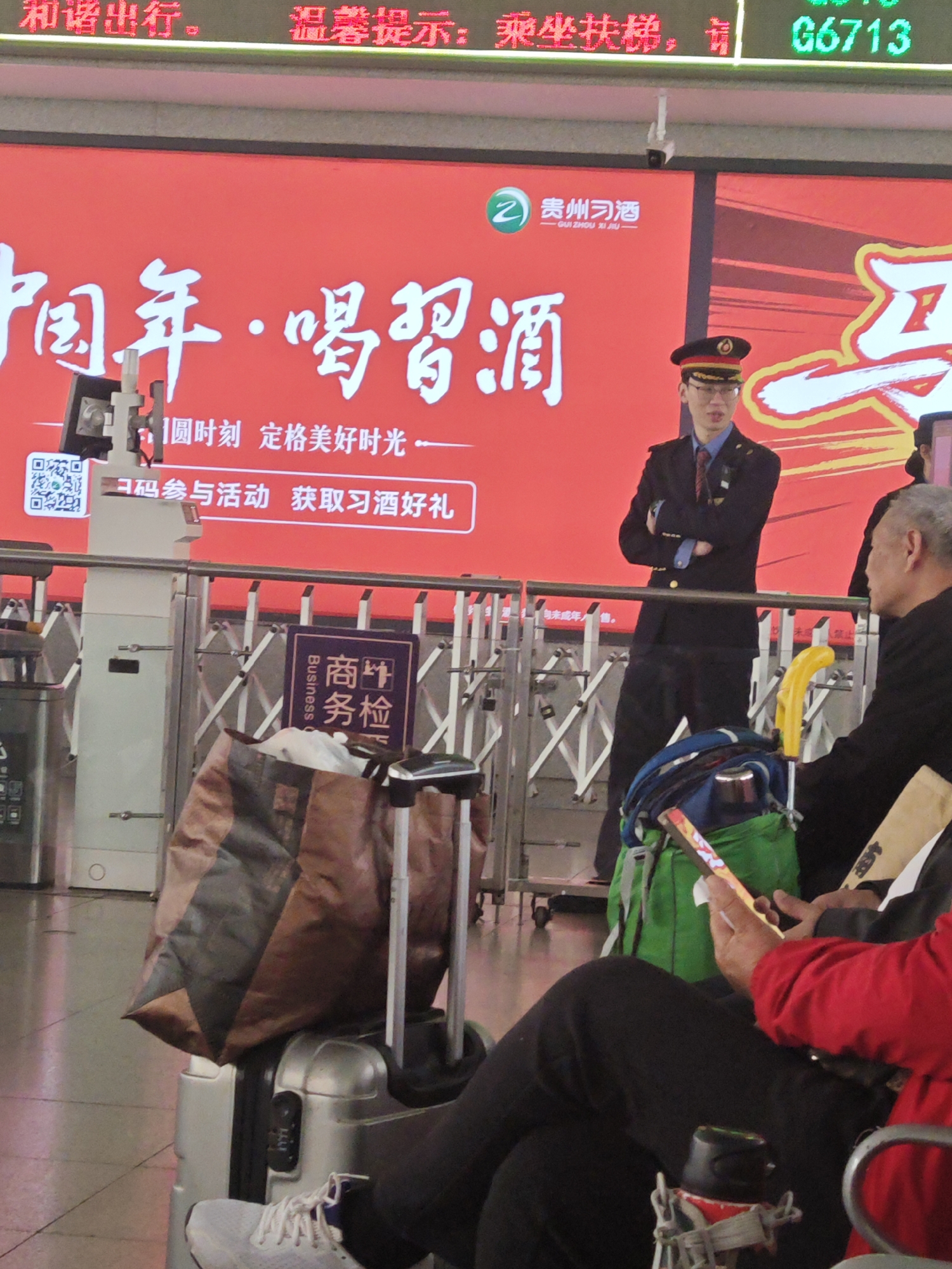This image depicts an indoor scene at a bustling transport hub, most likely a train station in China. Dominating the background is a large, vibrant red digital display screen. This screen features an advertisement for "贵州习酒" (Guizhou Xijiu) liquor, with the prominent slogan "中国年・喝习酒" (Chinese New Year · Drink Xijiu). Smaller text on the screen invites viewers to "扫码参与活动 获取习酒好礼" (Scan code to participate in the activity, get Xijiu gifts), next to a QR code. The top of the display shows public service messages like "和谐出行" (Harmonious travel) and "温馨提示:乘坐扶梯" (Warm reminder: take the escalator), along with the alphanumeric code "G6713," likely a train number.
In the foreground, a man in a dark uniform with a cap stands with his arms crossed, appearing to be an attendant or security personnel. To his right, an older man with grey hair is seated, partially obscured, facing away from the camera. Another person, wearing a red top and black pants, is seated in the immediate foreground, engrossed in a mobile phone. Luggage, including a silver suitcase, a large brown and black patterned bag, and a green backpack, is visible on the floor. A yellow cane handle protrudes from the green backpack. A vertical sign attached to a metal barrier reads "商务检" (Business Inspection), indicating a specific check-in or inspection point. The scene conveys a sense of people waiting in an organized, public transport environment.